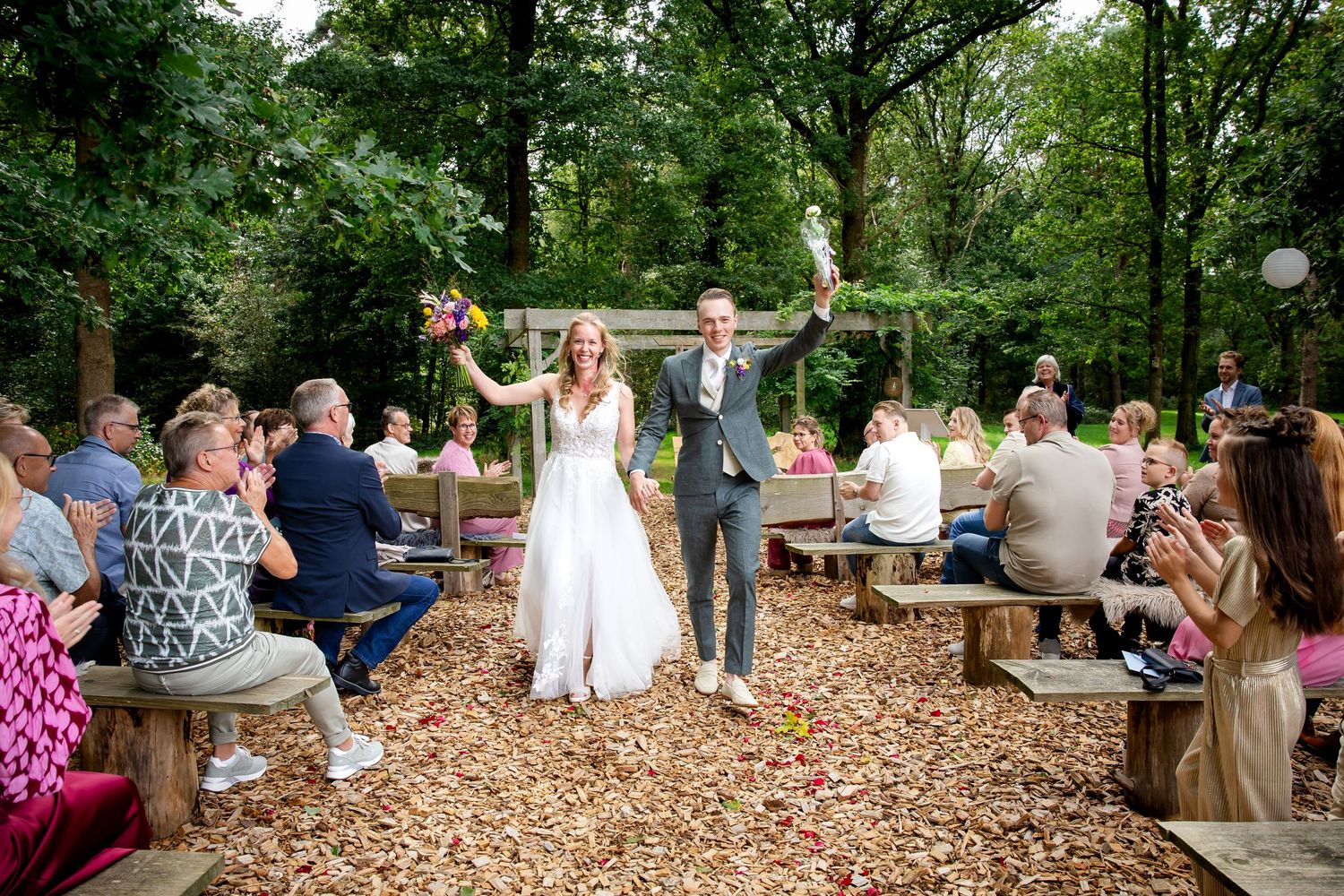 Outdoor wedding reception in wooded area with guests seated at picnic tables.