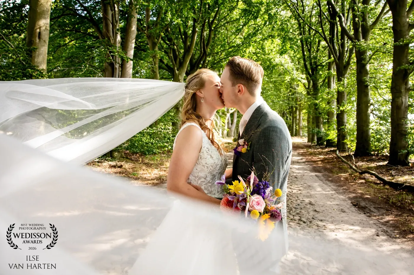 Bride and groom sharing a kiss on a forest path with veil flowing, award-winning wedding photo by Ilse van Harten.