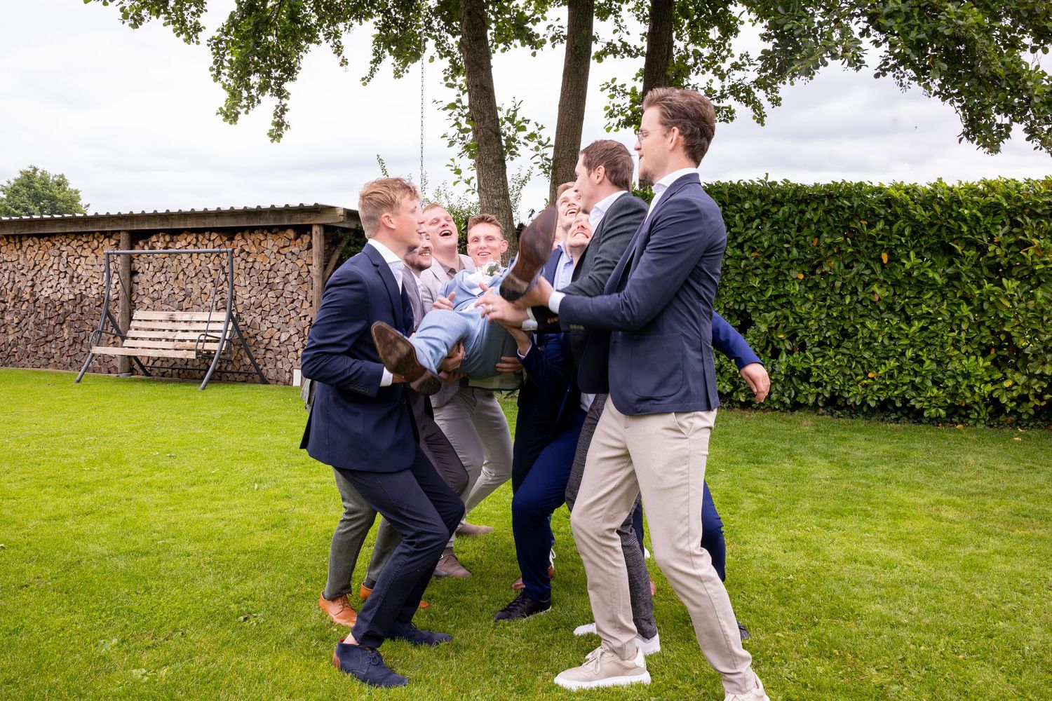 Group of men playfully wrestling and laughing on a green lawn with hedge backdrop.