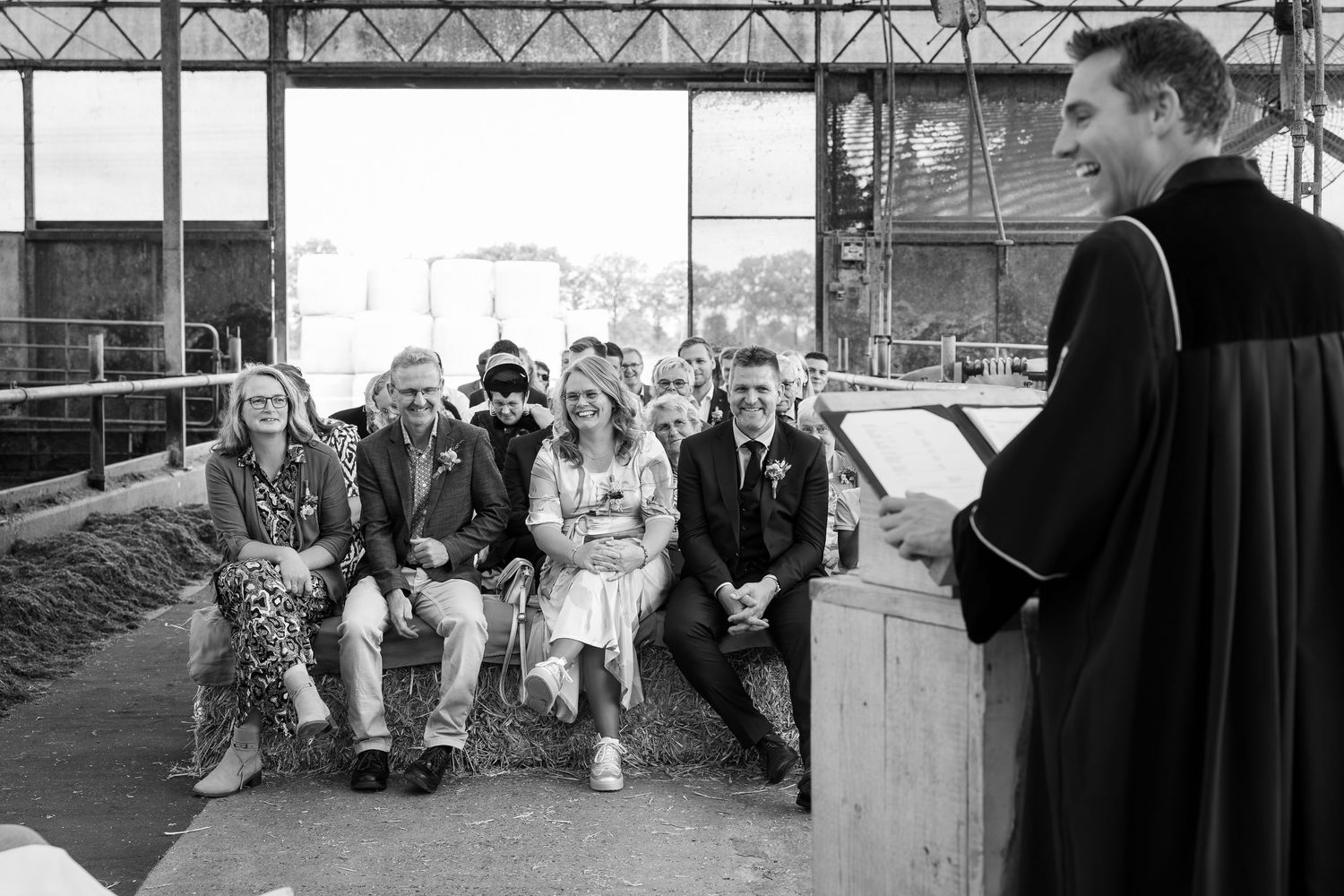 Black and white photo of a barn wedding ceremony with guests seated on rustic hay bales.