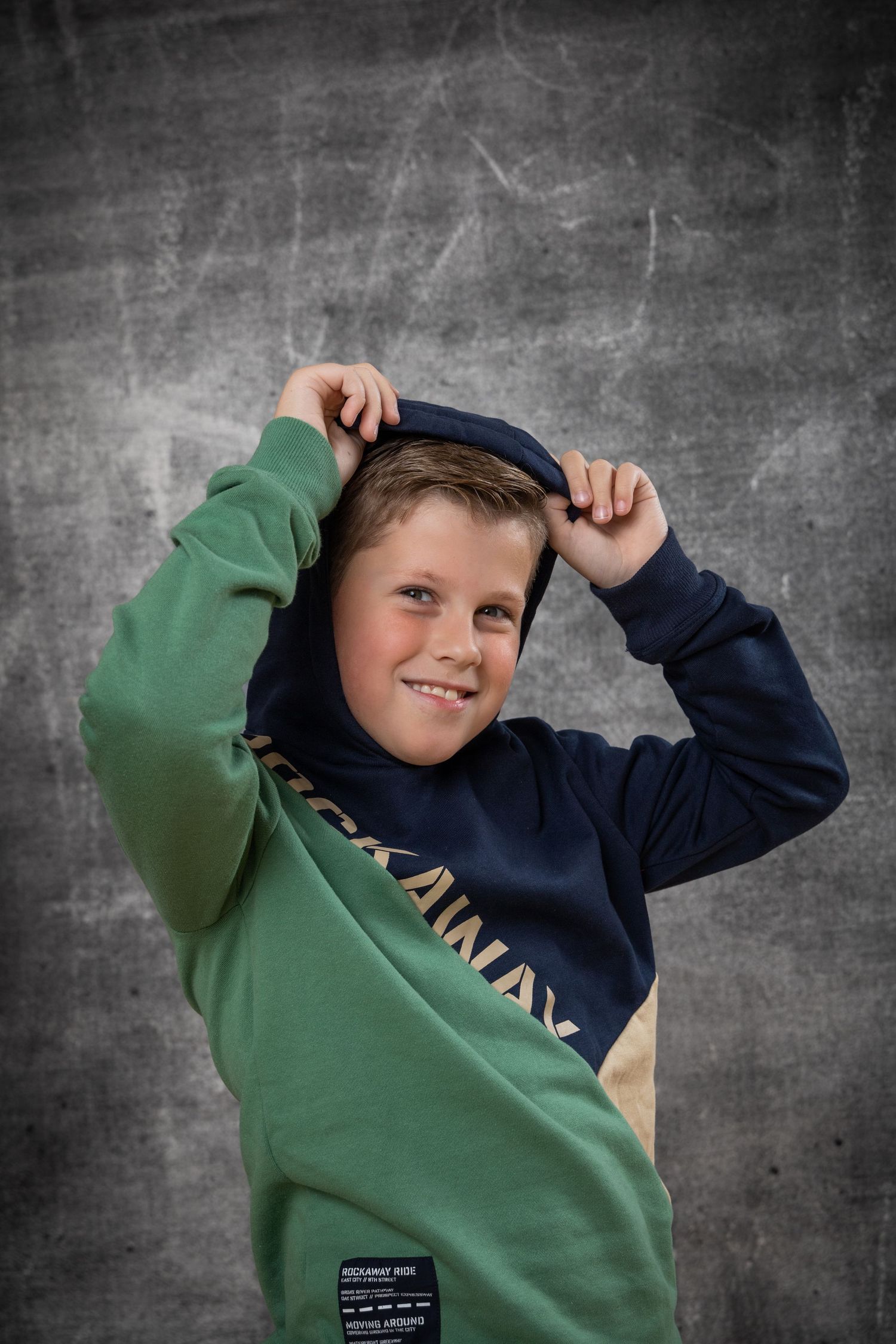 A young person in a two-toned navy and green sweatshirt poses playfully against a dark textured background.