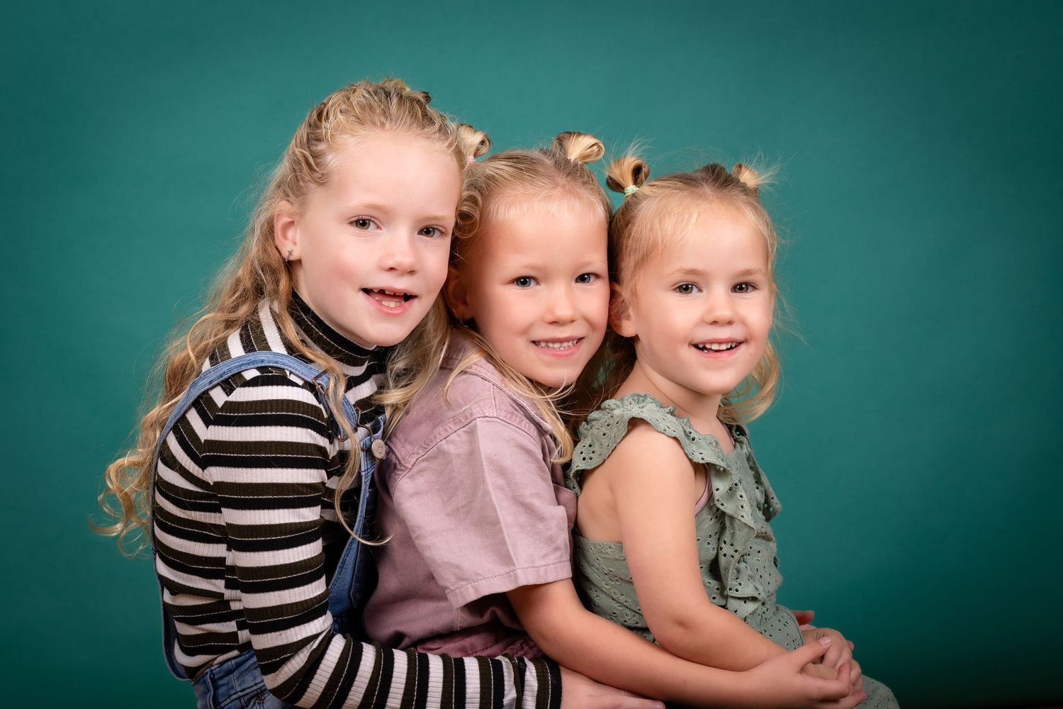 Three children in casual wear pose together against a teal studio backdrop.