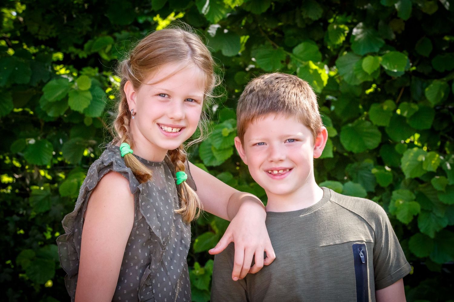 Two people smile together outdoors with lush green foliage in the background.