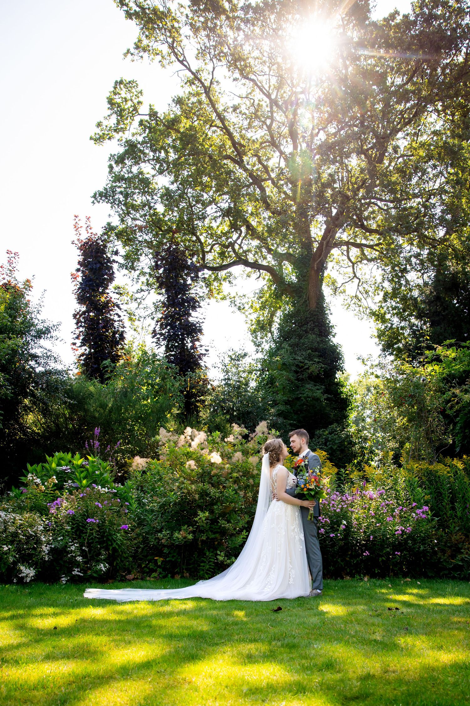A wedding couple embraces in a lush garden setting with dramatic sunlight streaming through tall trees.
