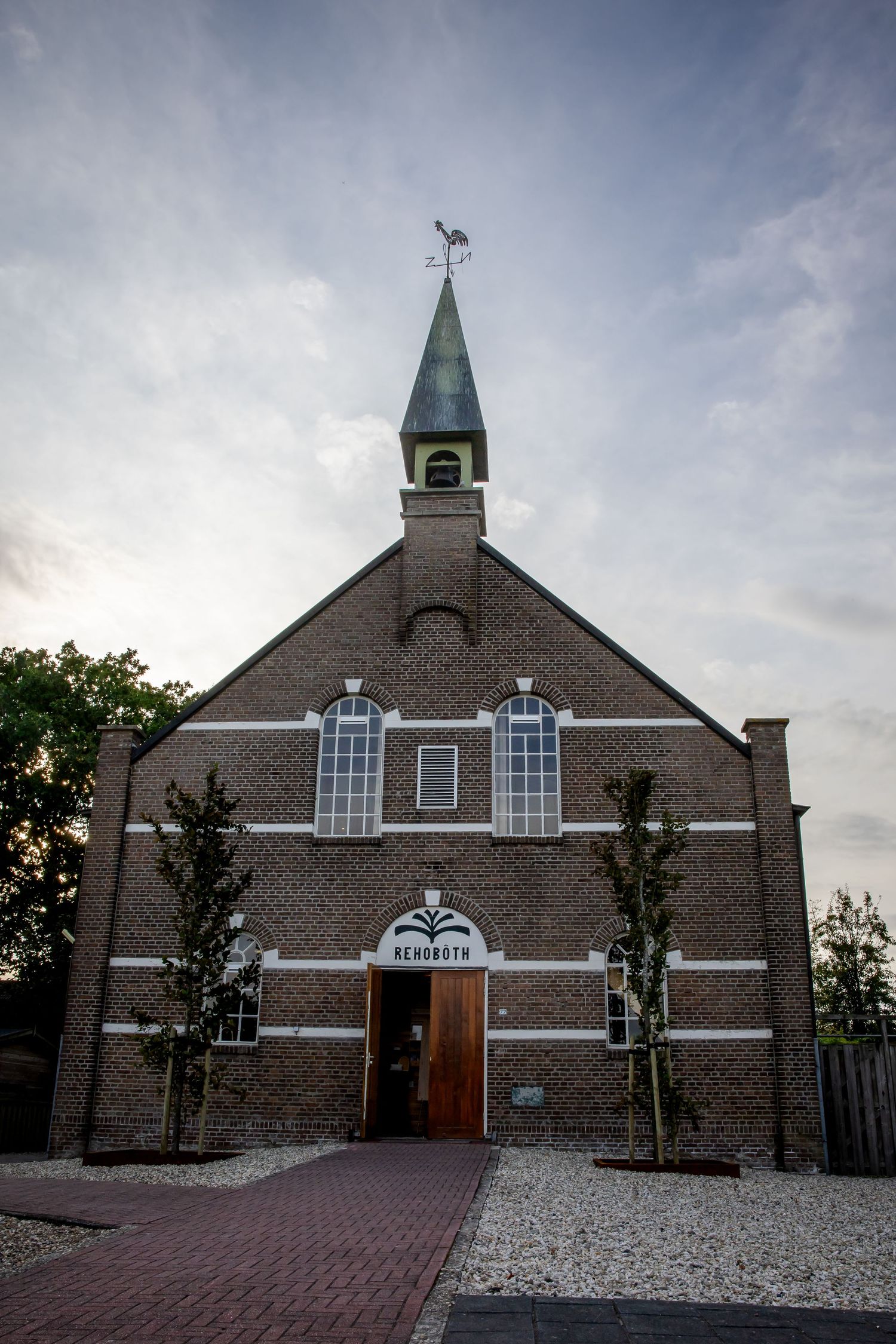 Buitenaanzicht van een bakstenen kerkgebouw met een groene torenspits en witte ramen in de schemering.