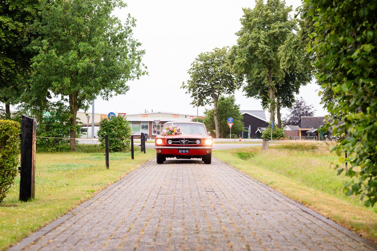 A red vintage car sits parked at the end of a tree-lined brick path.