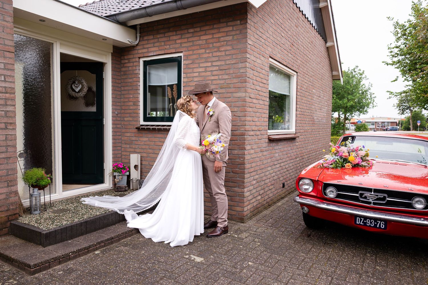 A wedding scene unfolds outside a brick house with a red vintage car in the background.