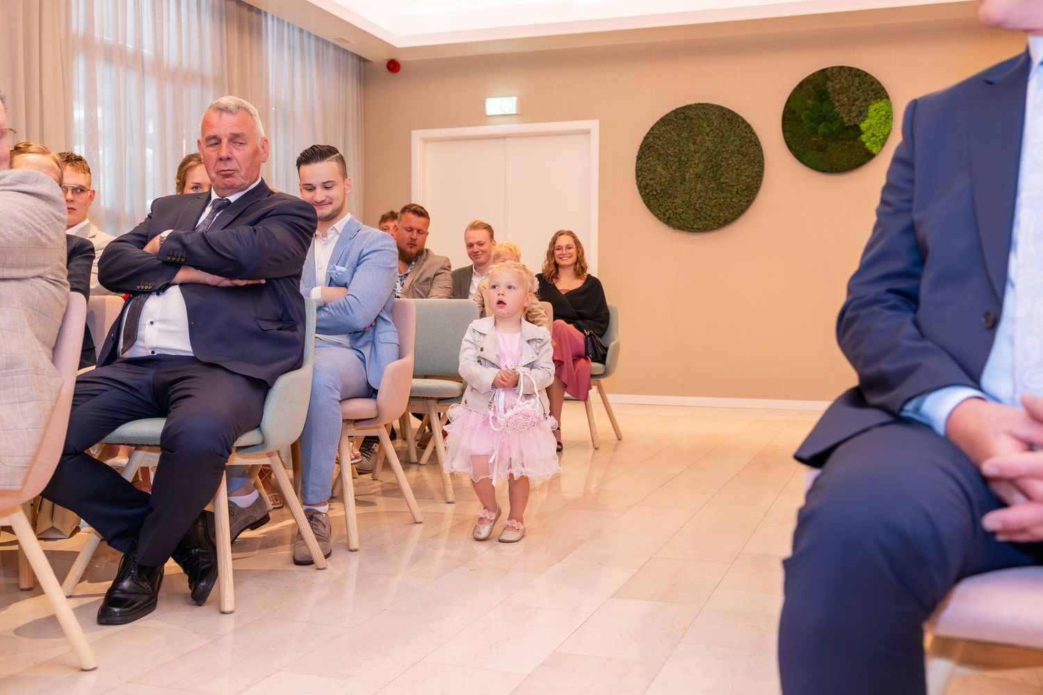 Wedding guests seated on wooden chairs in a modern room with moss wall art.