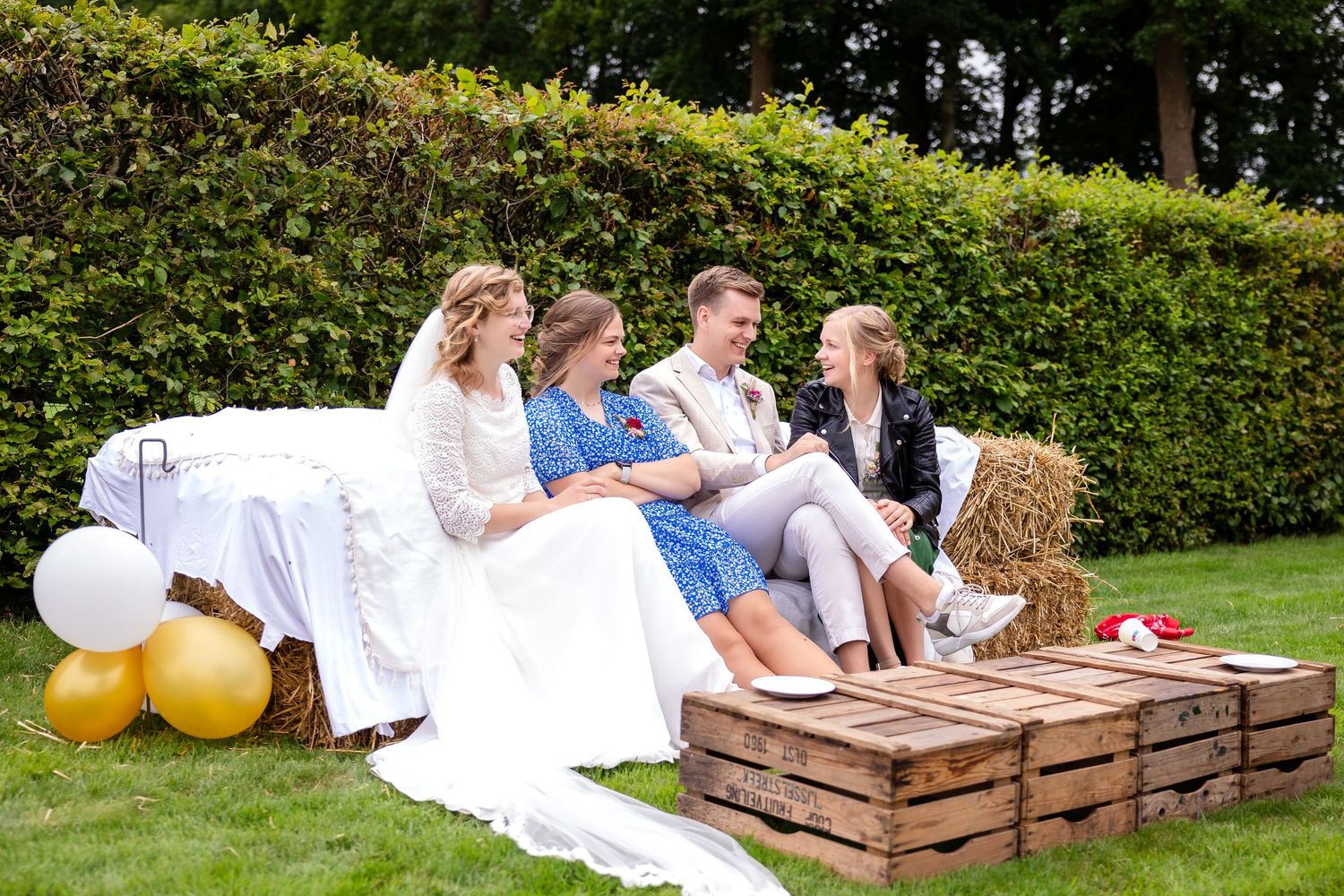 Outdoor wedding party sitting on rustic wooden crates with yellow balloons in a garden setting.