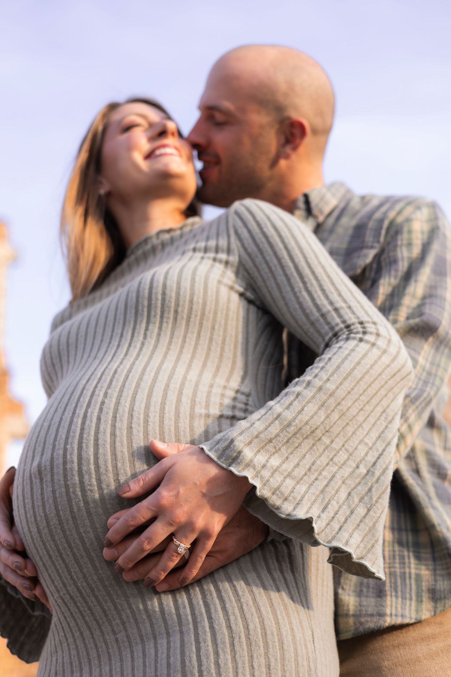 Expectant couple embracing outdoors, man kissing pregnant woman as she smiles during maternity session.
