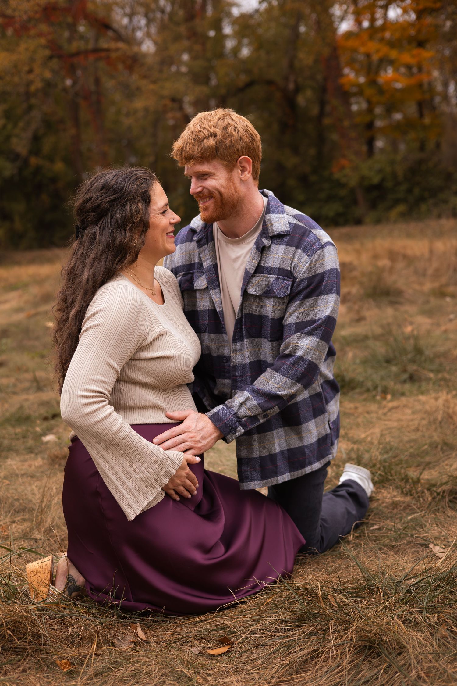 Smiling pregnant couple sit together on autumn grass, man in plaid shirt embracing woman in burgundy skirt.
