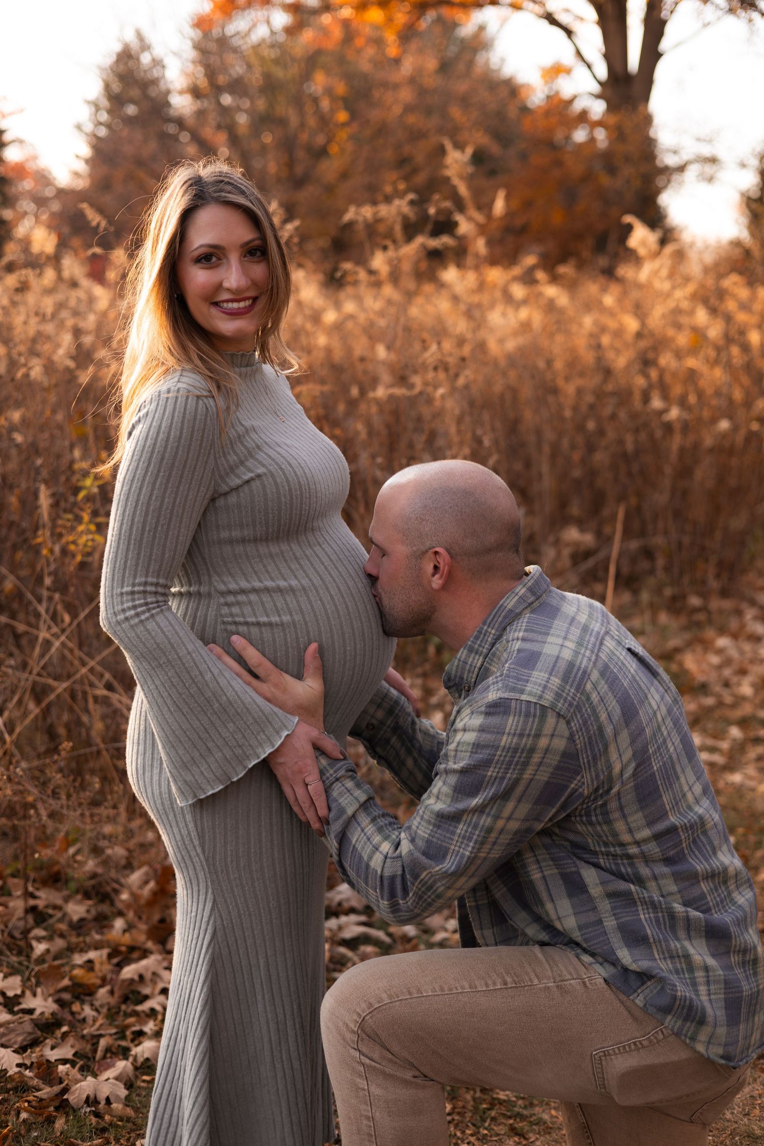 Man kneeling to kiss pregnant woman's baby bump during golden hour fall maternity photo session.