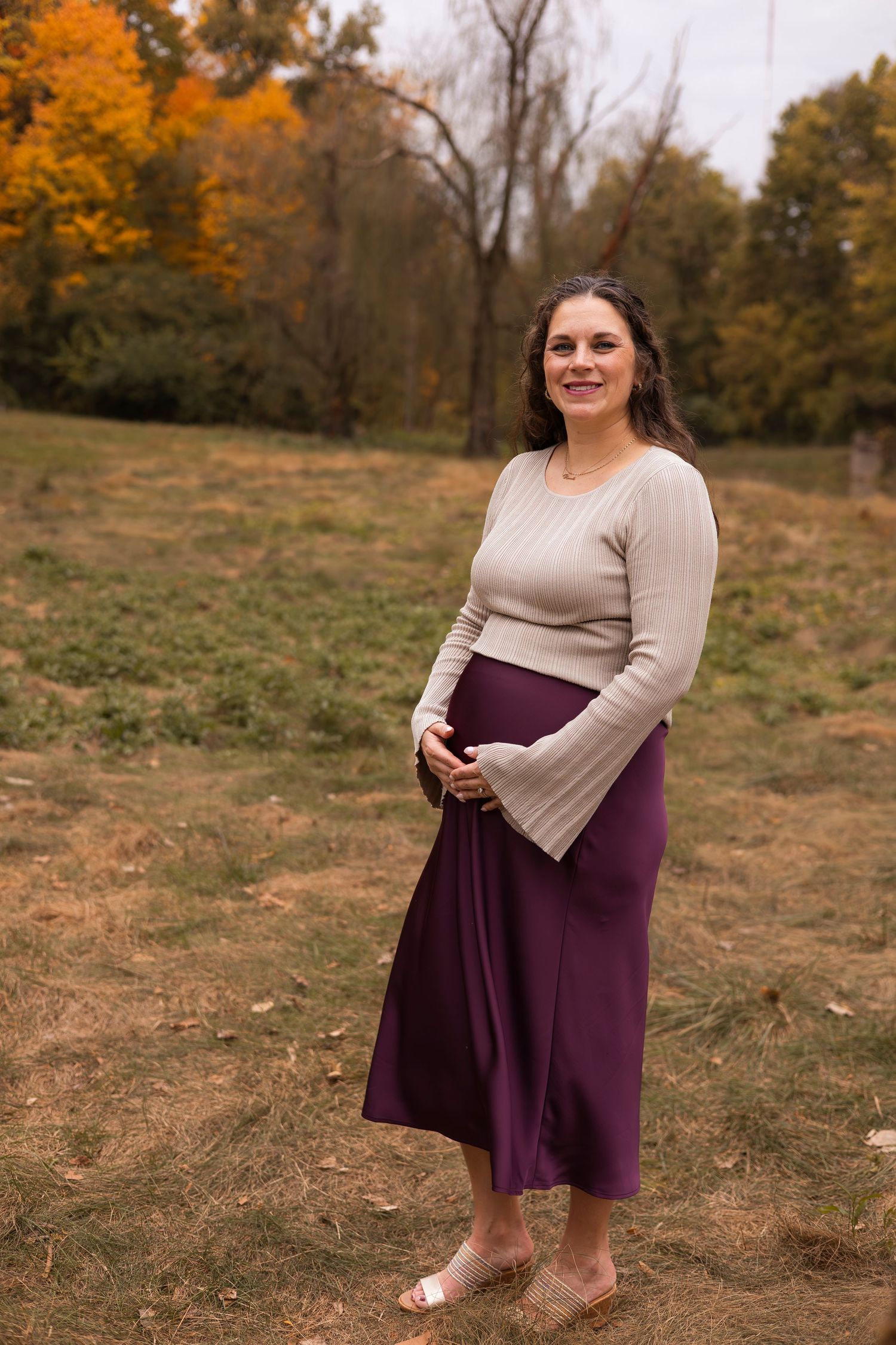 Smiling pregnant woman in beige sweater and burgundy skirt cradles baby bump in colorful autumn park setting.