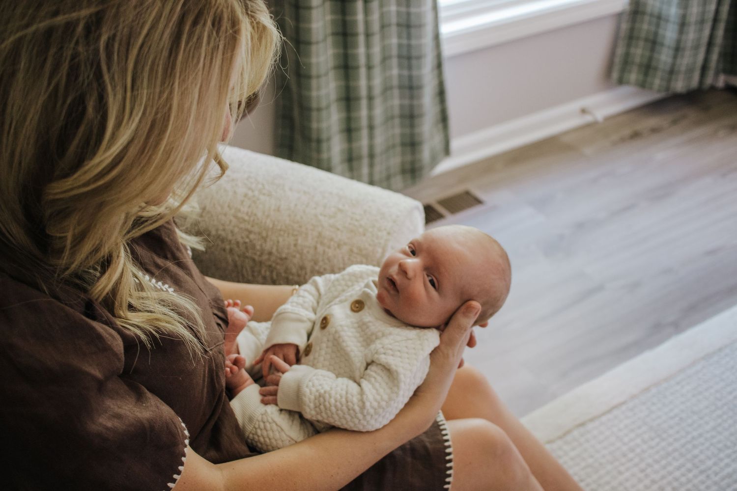 Blonde mother sitting in chair lovingly gazing at newborn in white knit outfit.