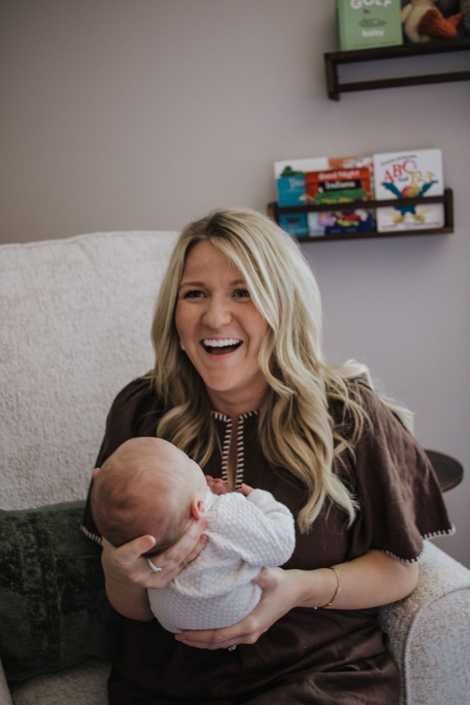 Joyful blonde mother laughing while holding sleeping newborn baby in nursery chair.