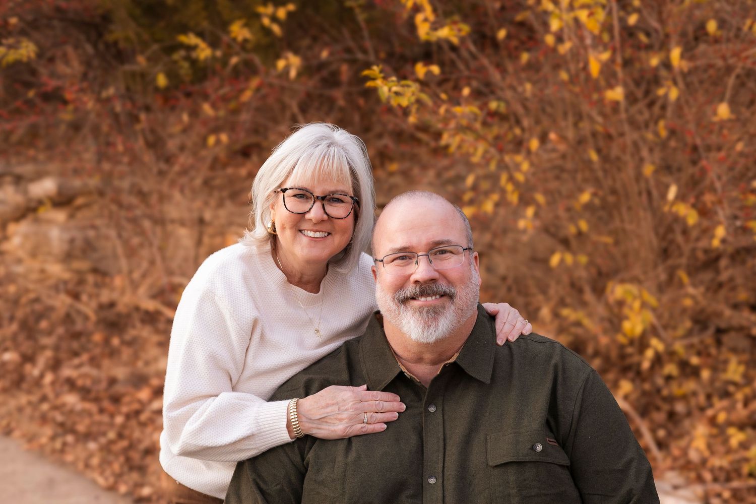 Older couple smiling warmly outdoors with golden autumn leaves in background.
