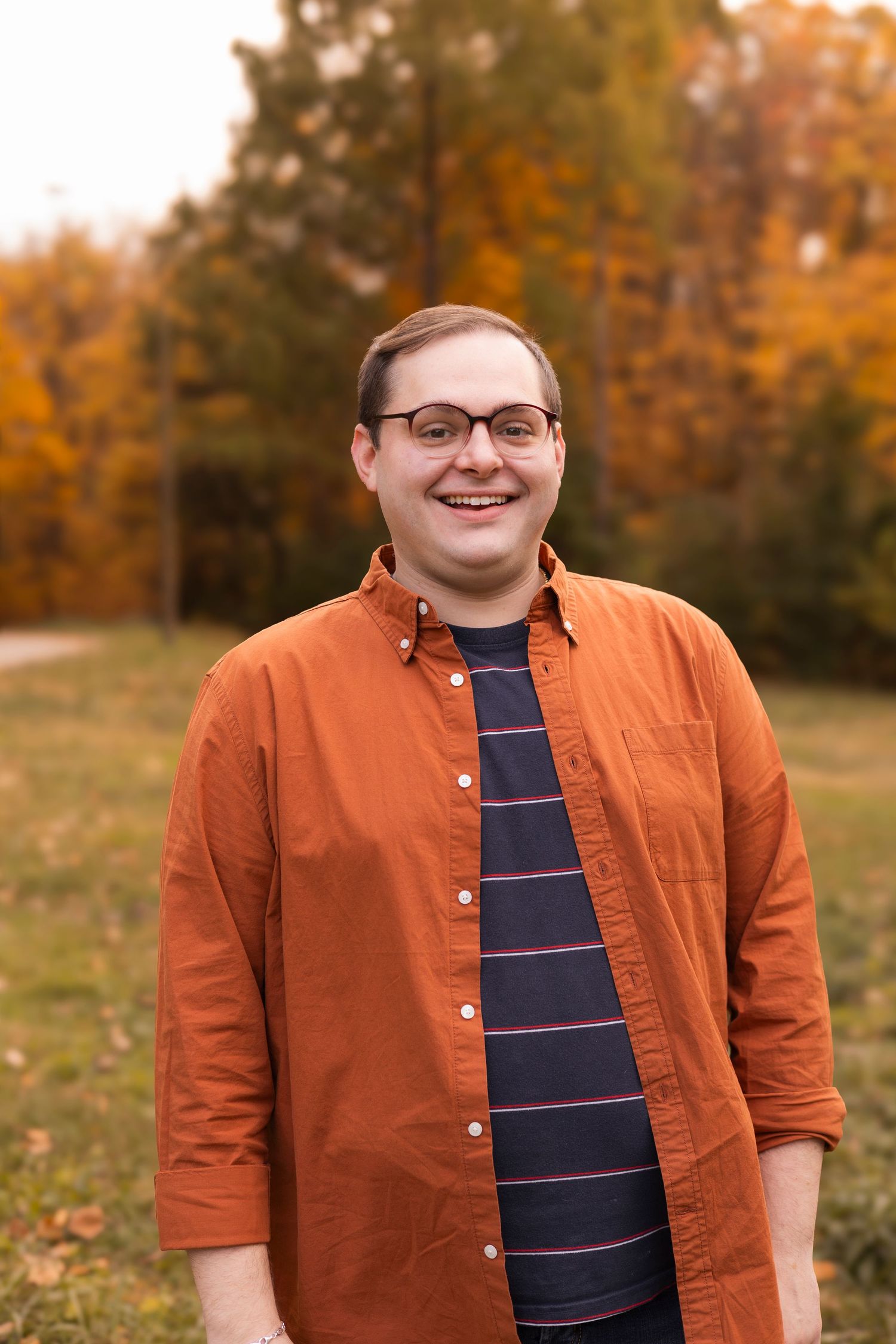 Smiling young man in orange shirt and glasses standing outdoors among autumn foliage.