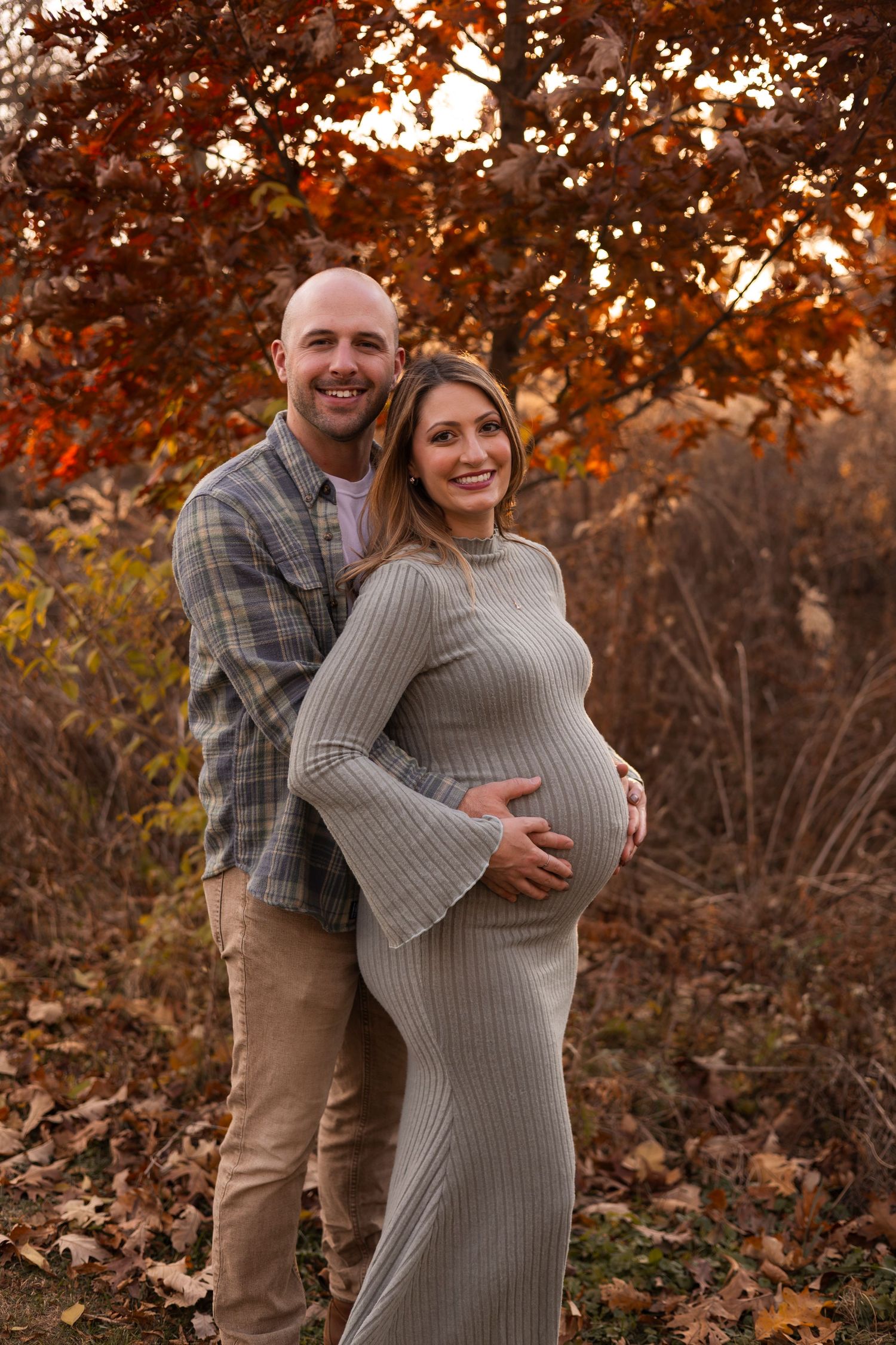 Happy expecting couple embracing under red autumn foliage during romantic fall maternity photo session.