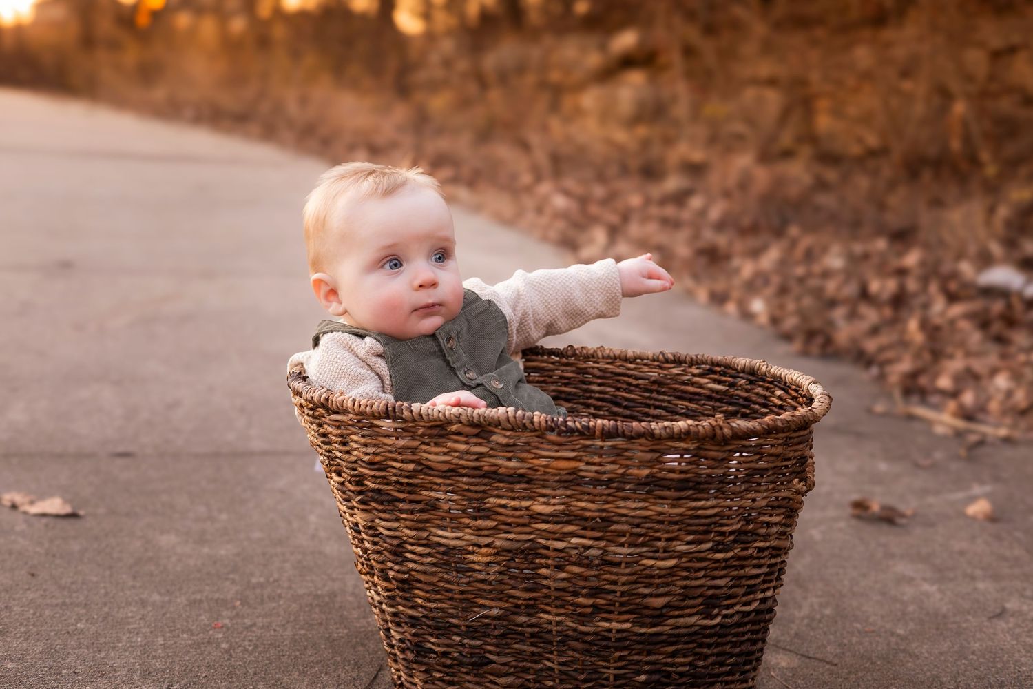 Curious baby leaning over edge of wicker basket on path with fall leaves.