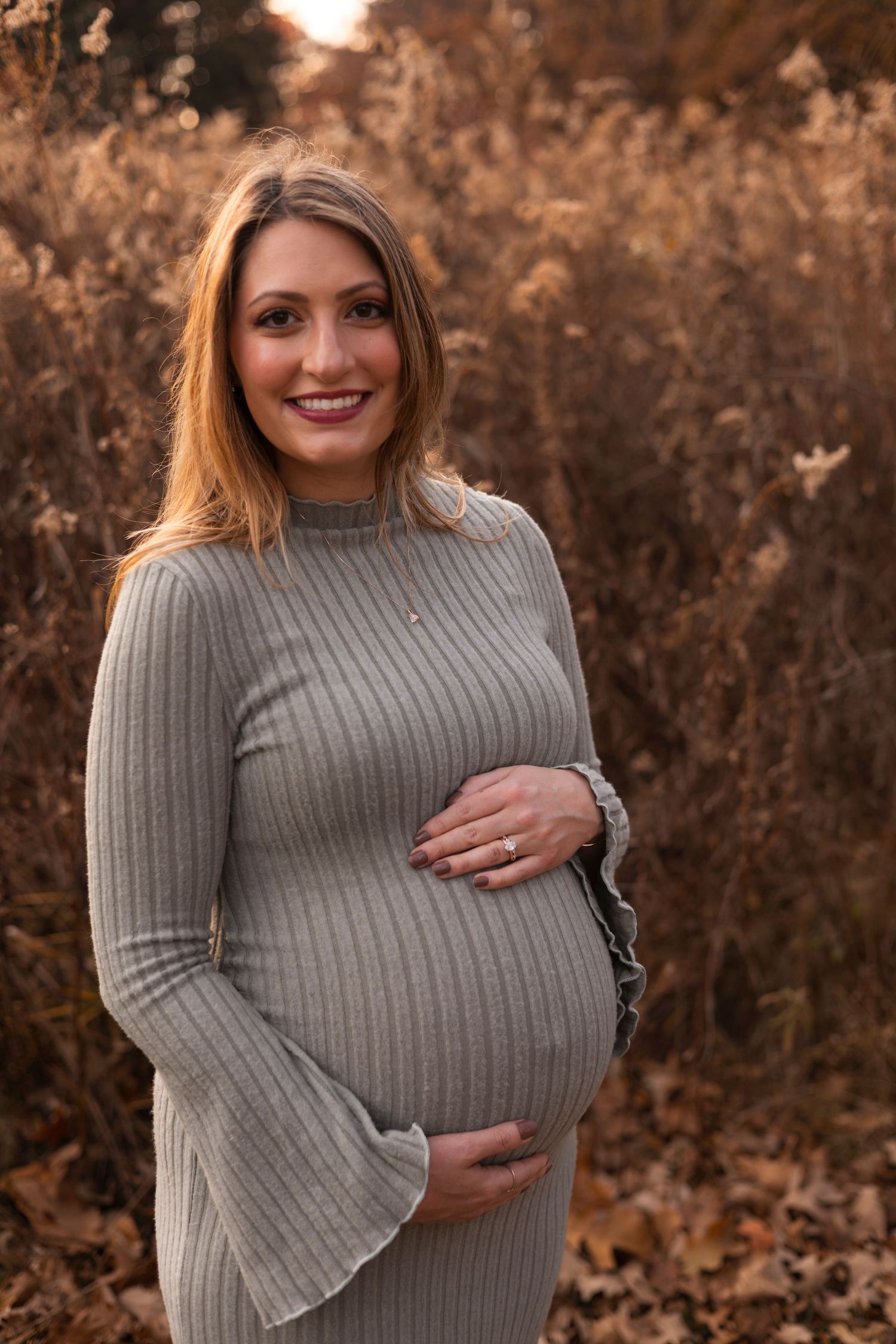 Smiling pregnant woman in gray ribbed dress cradling baby bump during autumn outdoor maternity session.