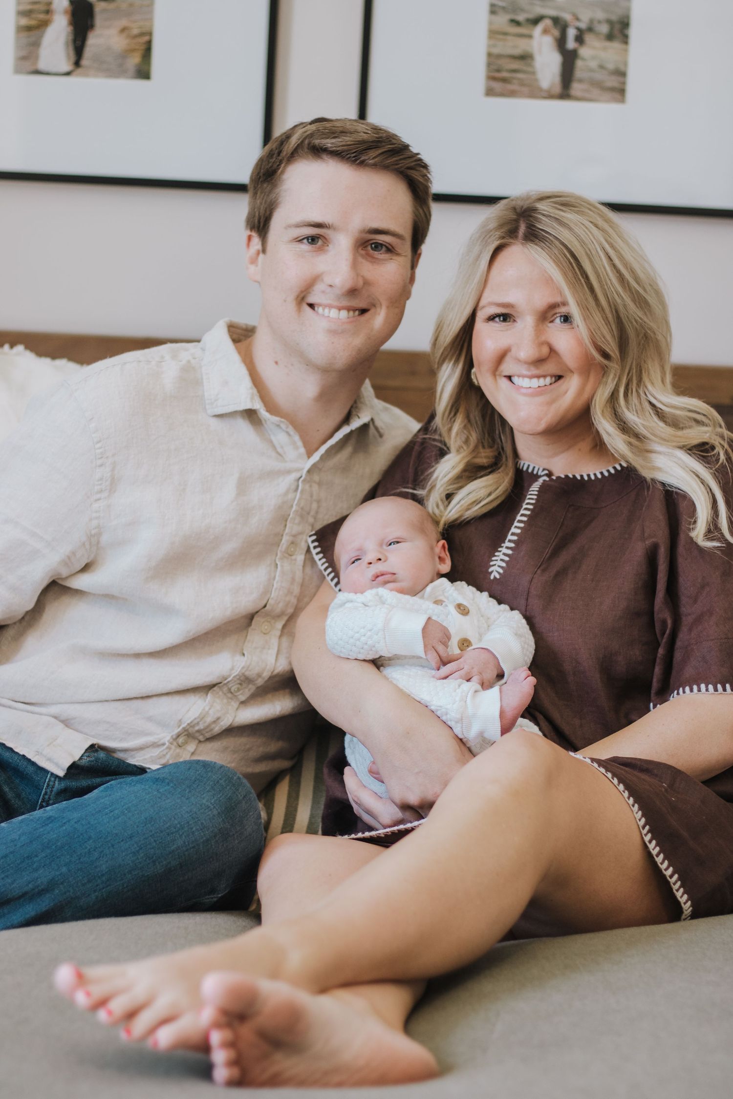 A smiling young couple sitting on a couch indoors, proudly holding their newborn baby.
