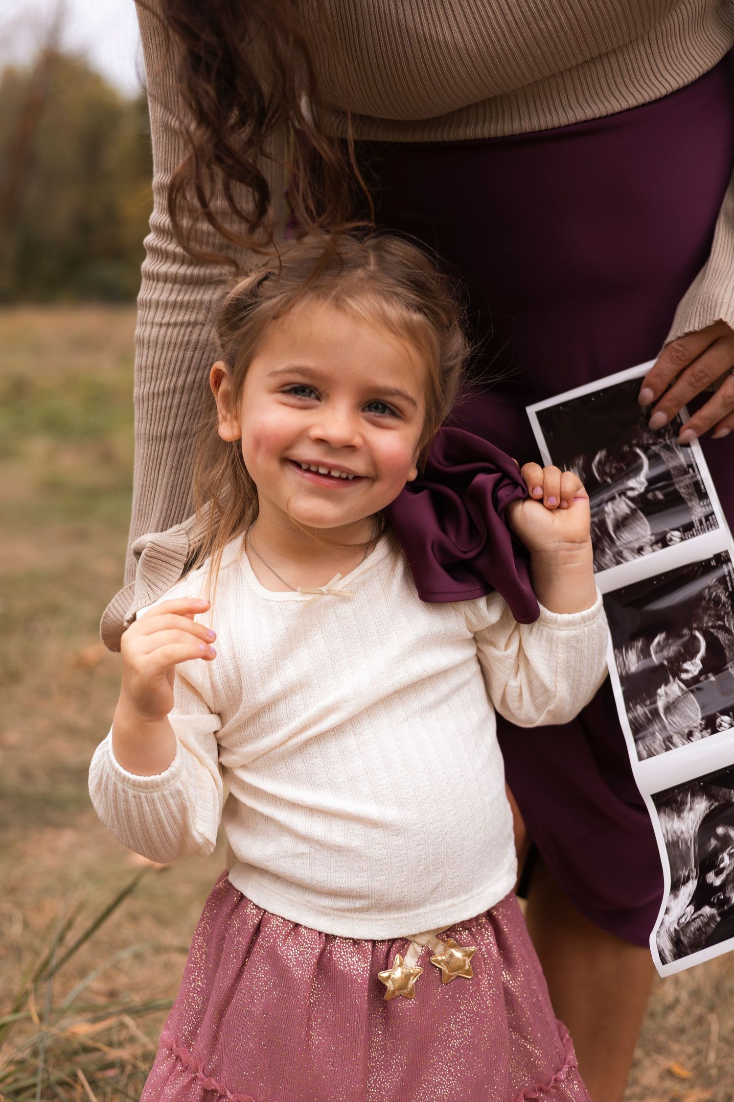 Smiling toddler girl in pink skirt holds ultrasound photo beside her pregnant mothers burgundy skirt outdoors.