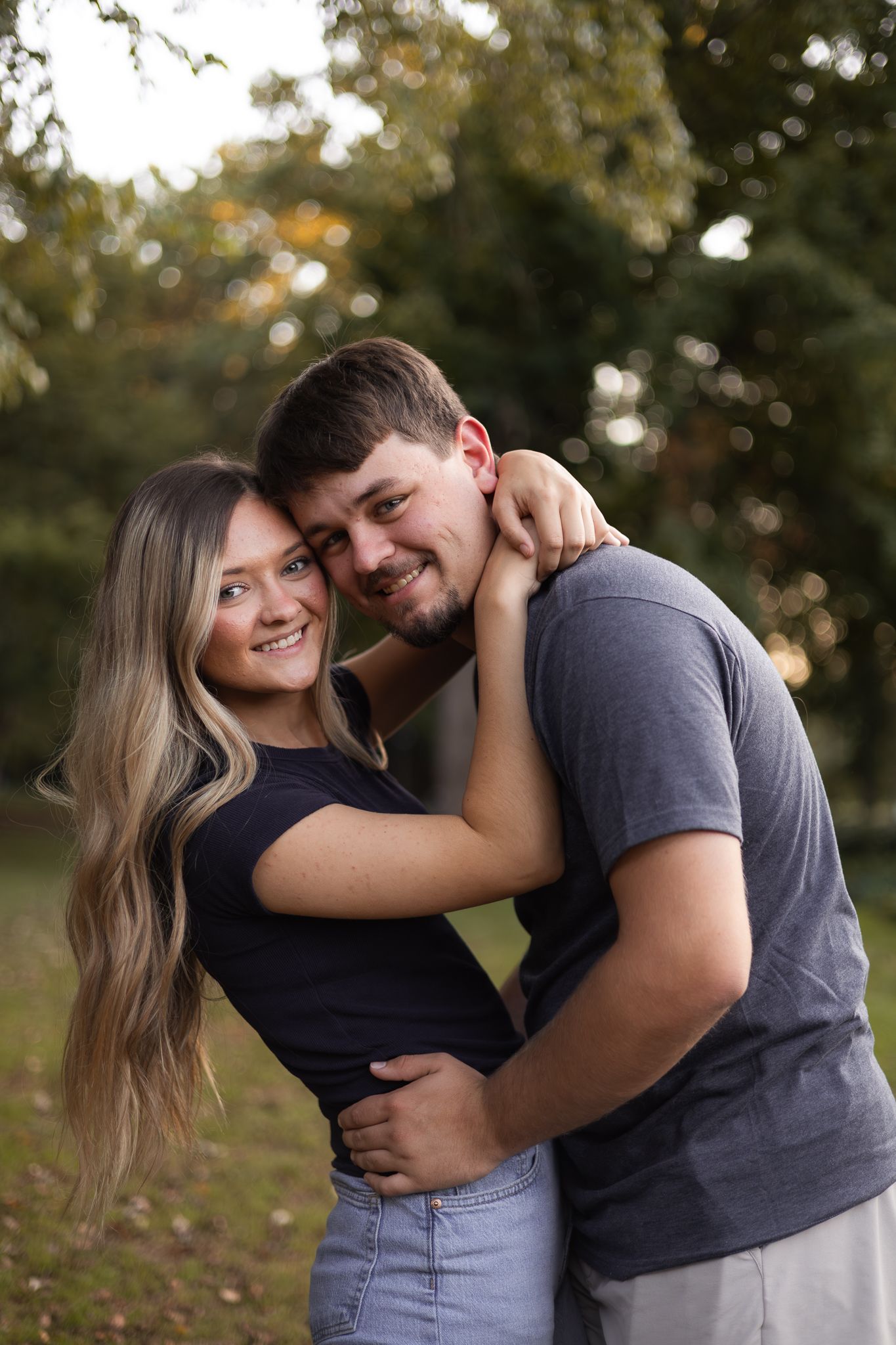 A smiling young couple cuddling together in a lush green outdoor setting during golden hour.
