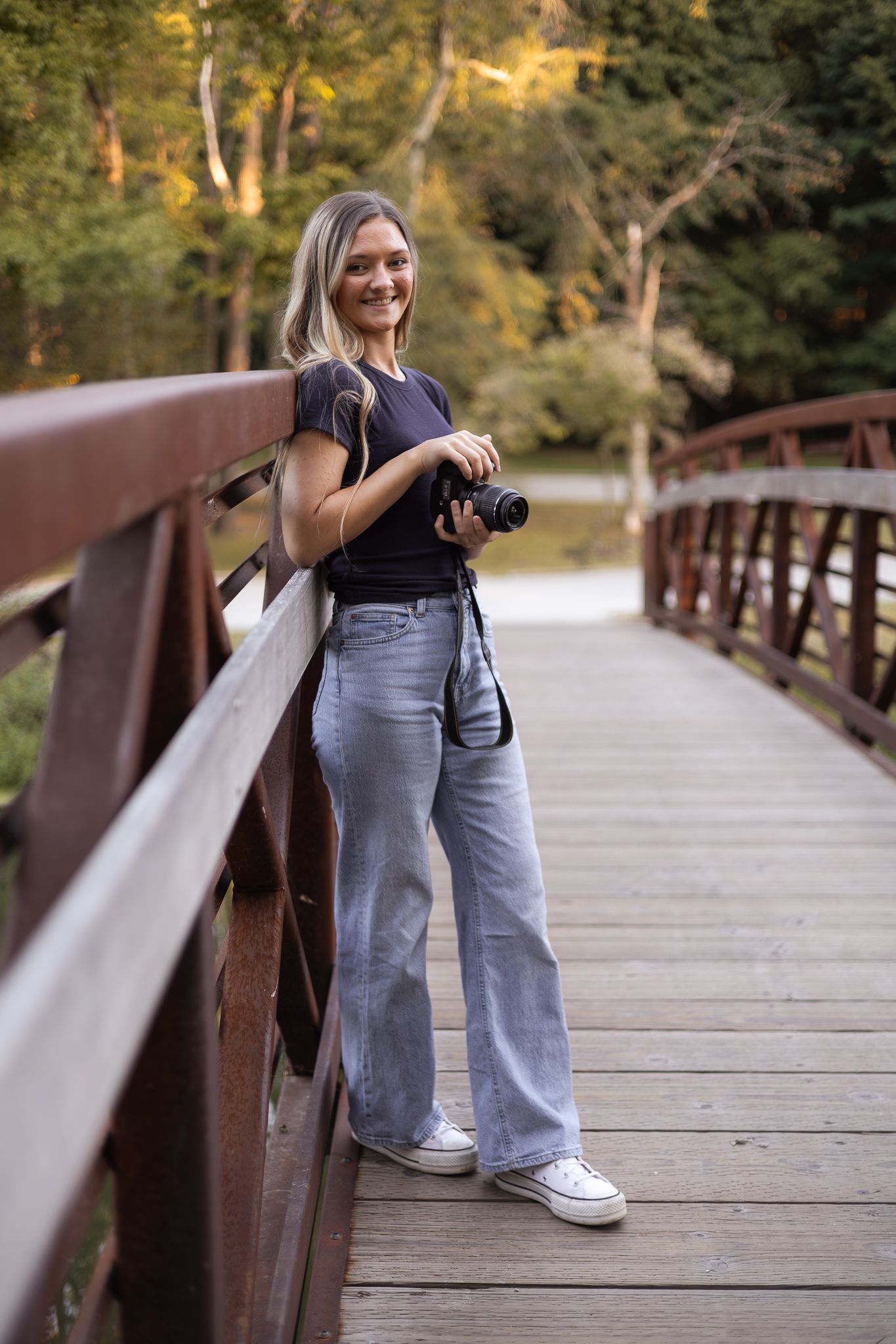 Young blonde woman holding camera on wooden bridge for outdoor portrait session.