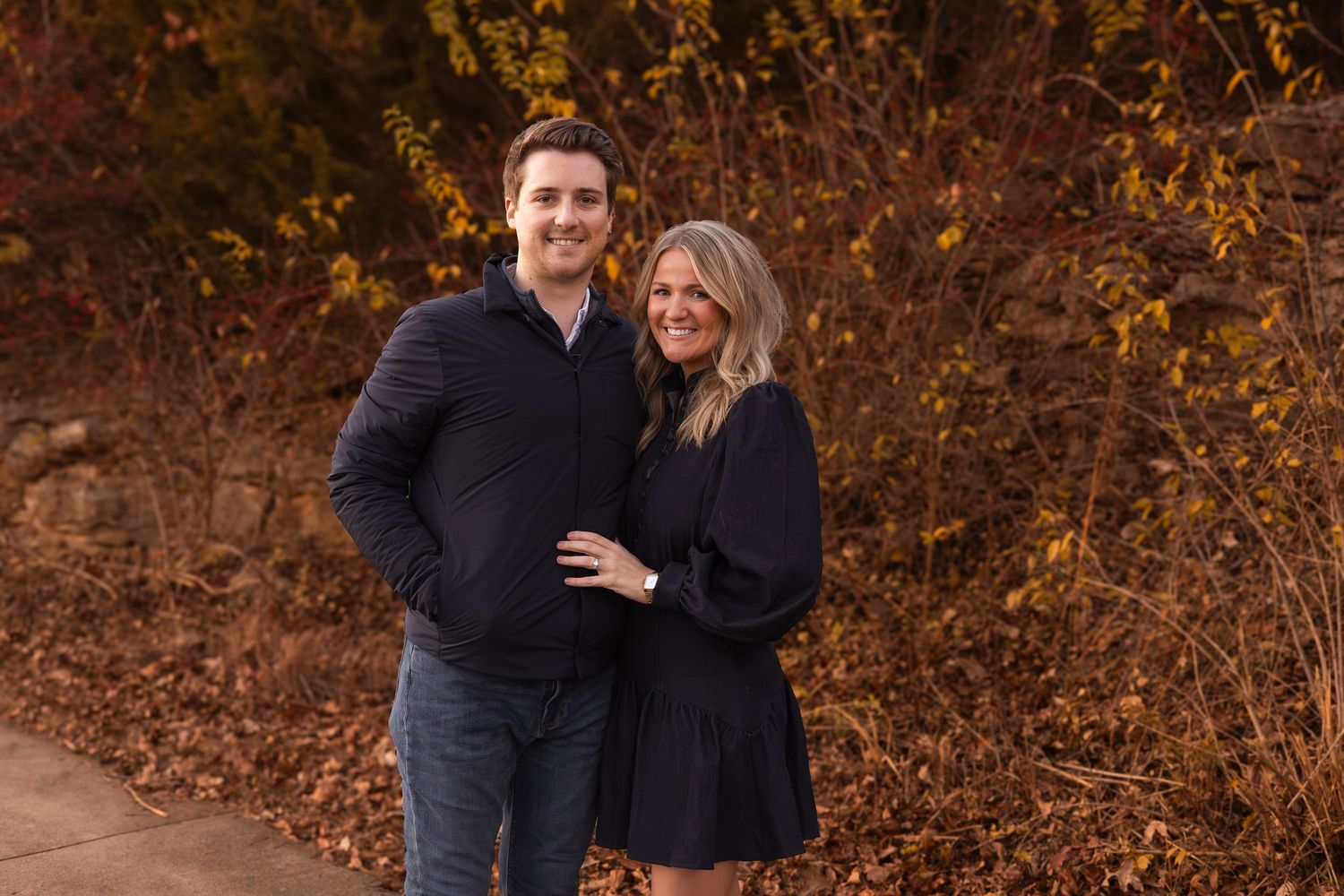 Smiling couple poses together on a path surrounded by rich autumn foliage during an outdoor portrait.