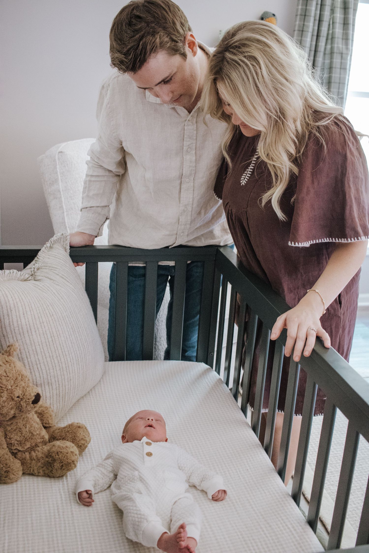 New parents smiling down at newborn baby lying in dark wood crib with teddy bear.