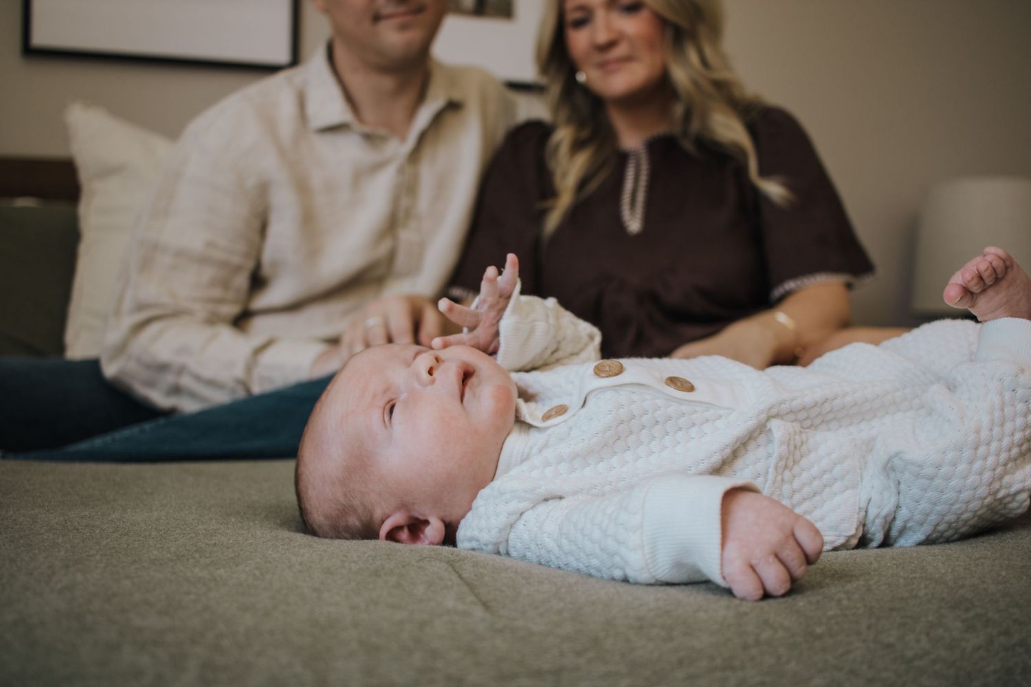 Newborn baby lies on a gray couch while parents smile in the background.