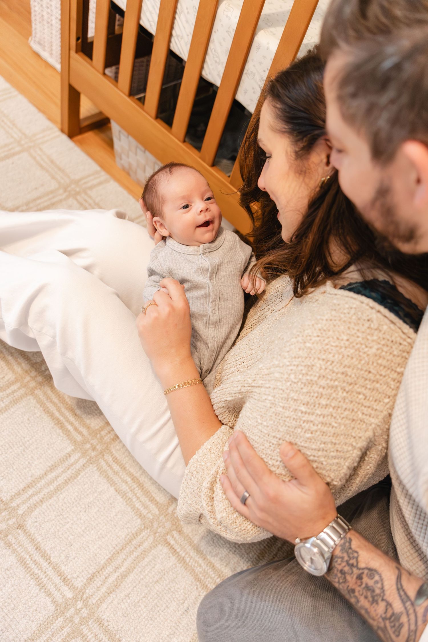 Parents sitting on floor with newborn baby by wooden crib, sharing a tender family moment.