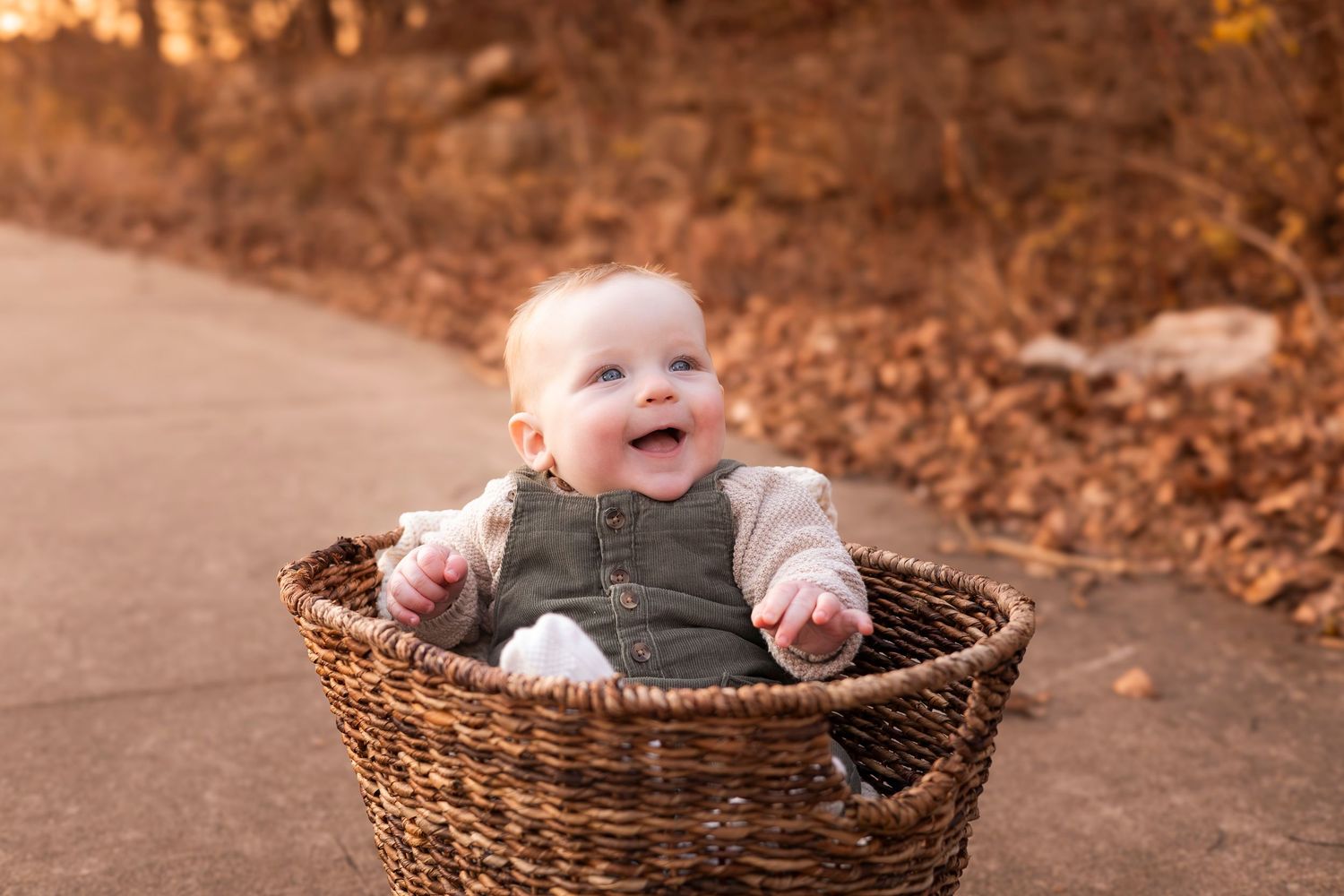 Smiling baby sitting in wicker basket on outdoor path with autumn foliage.
