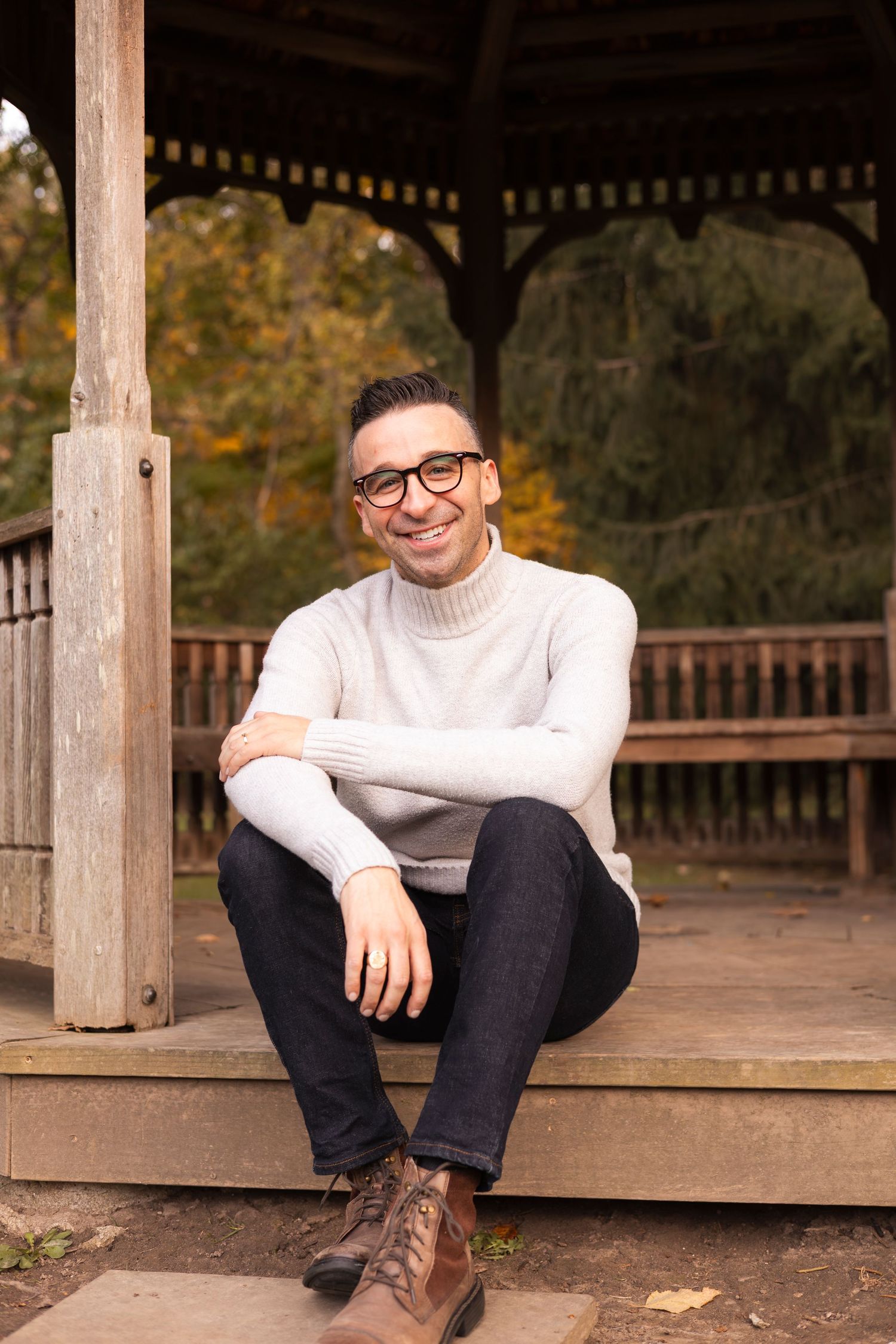 Smiling man in white turtleneck sweater sitting relaxed under a wooden gazebo in autumn park.