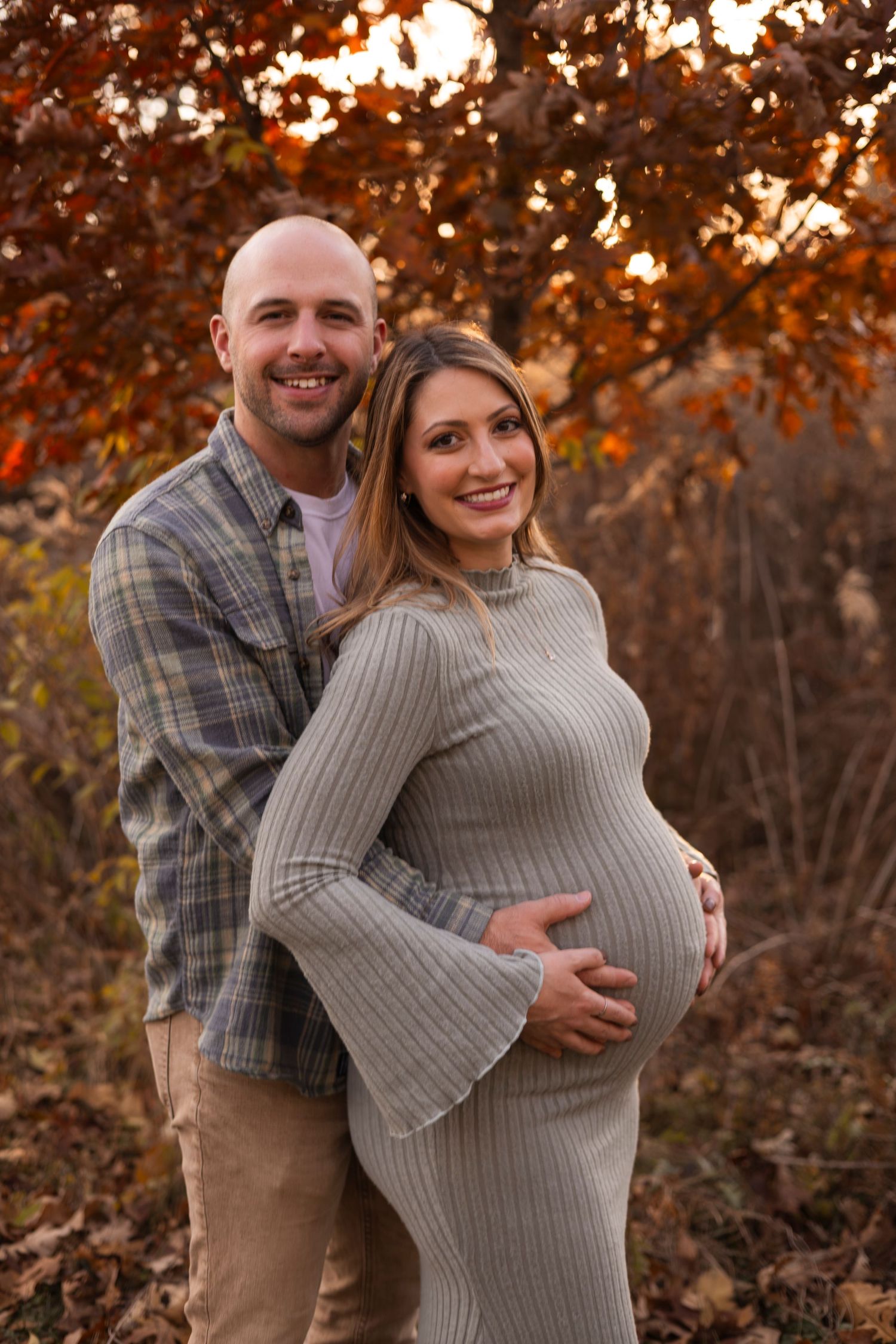 Couple poses outdoors during fall maternity photo session, man embracing pregnant woman in gray ribbed dress among autumn foliage.
