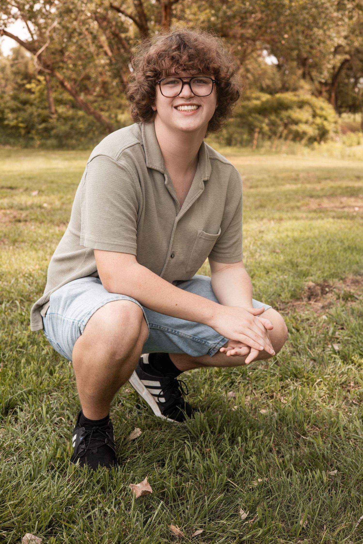 Teenage boy with curly hair and glasses crouching on grass in park setting.