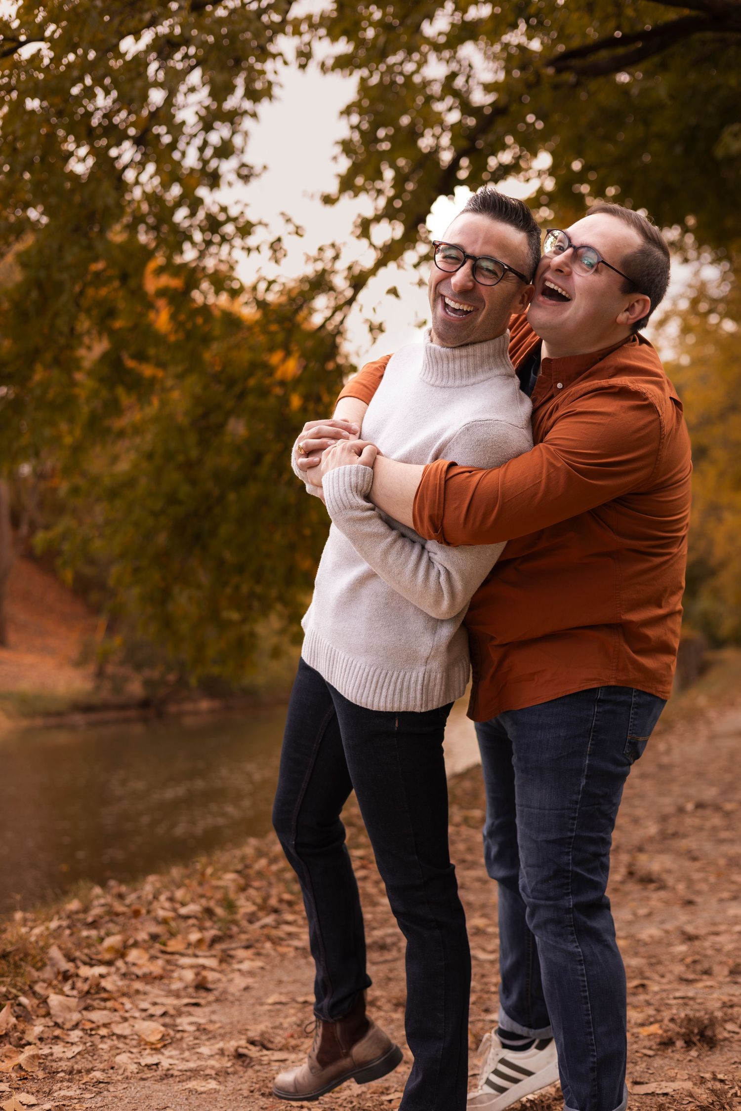 Two men laughing and hugging outdoors near a river in autumn.