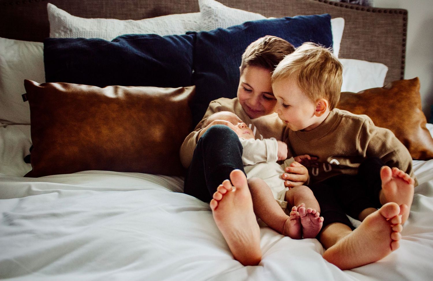 Two young brothers cuddle together on a white bed with soft leather and navy pillows in the background.