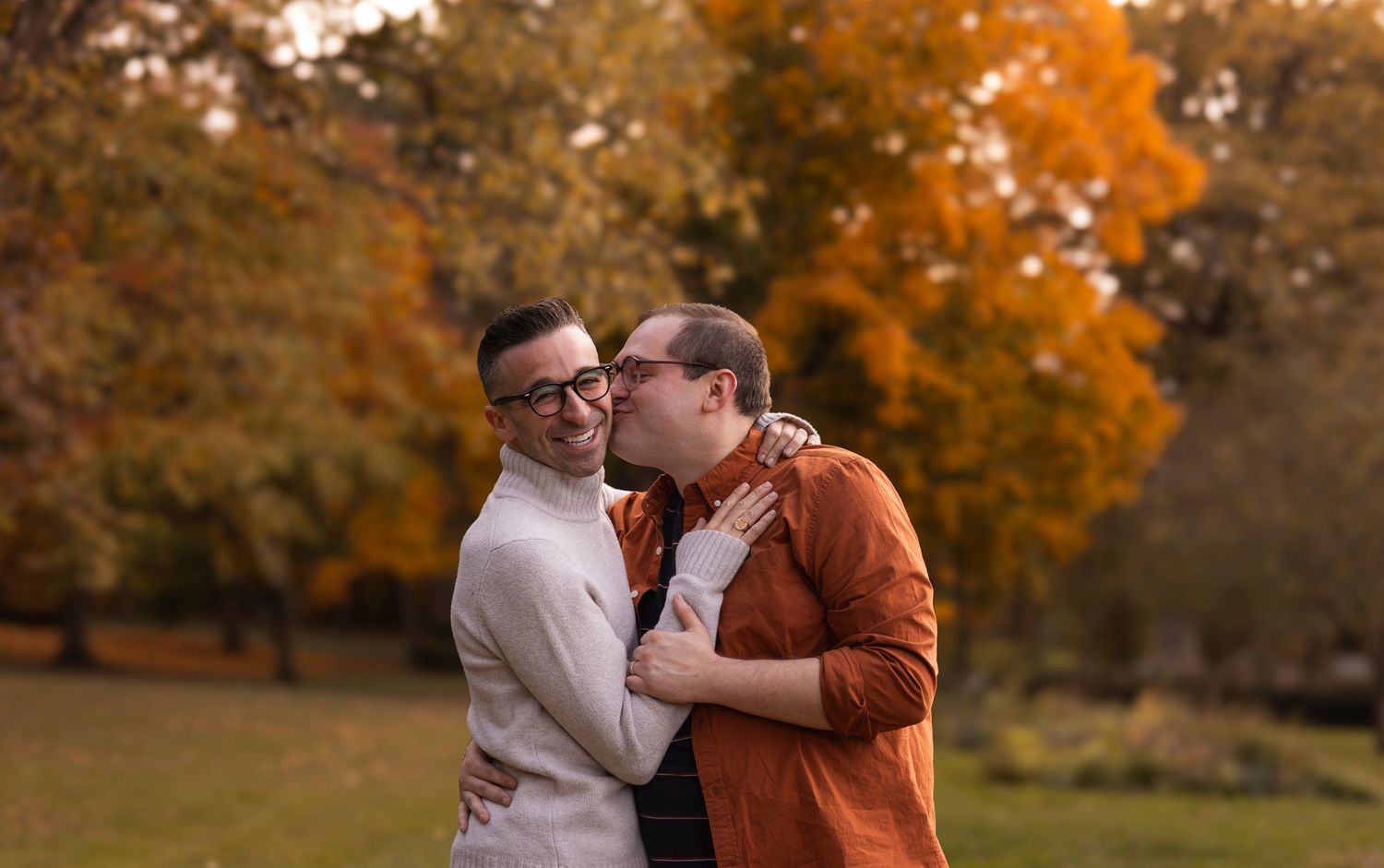 Two men sharing a kiss in a colorful autumn park setting.