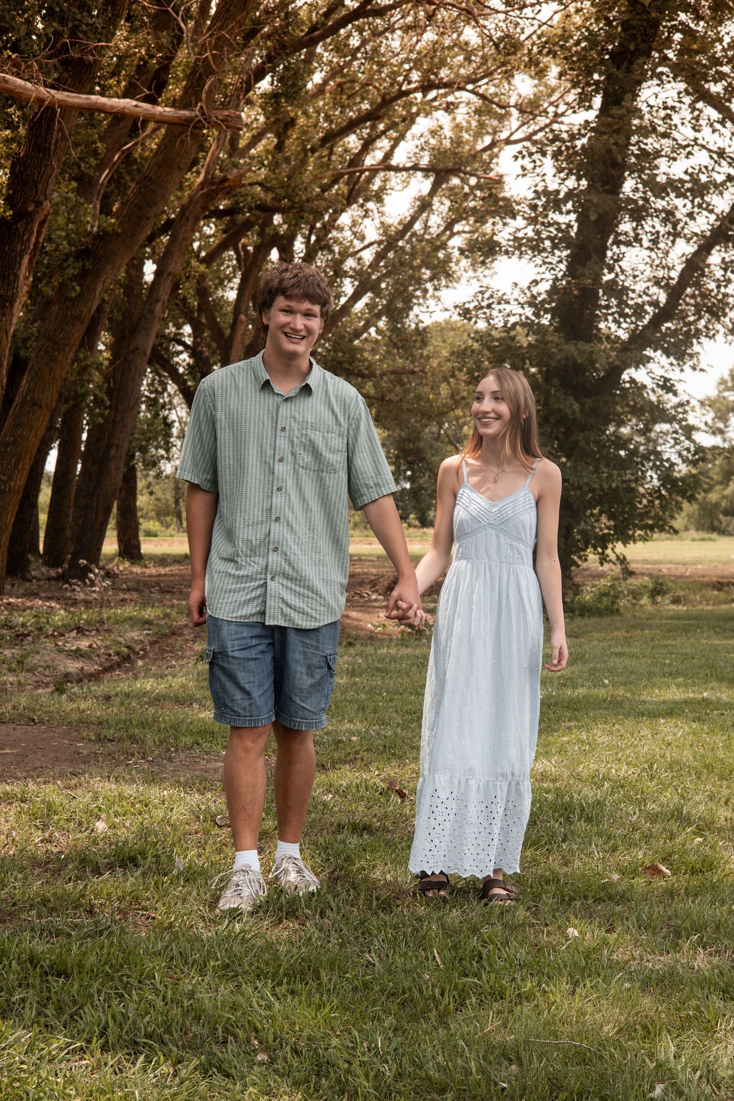 Young couple holding hands and walking through a scenic tree-lined grassy park.