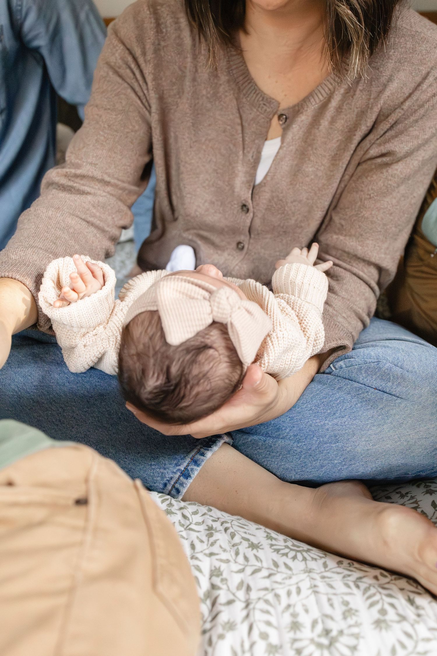 Mother holds sleeping newborn baby in knit outfit and pink bow headband on her lap.