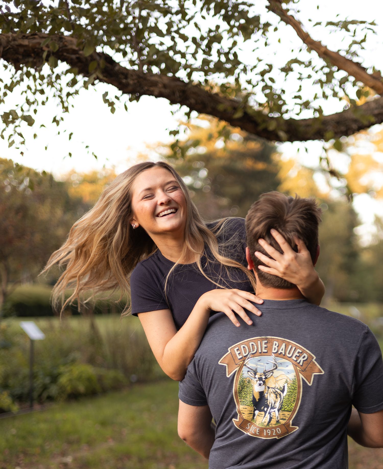 A joyful young woman laughing as her partner lifts her under a tree in a sunny park.