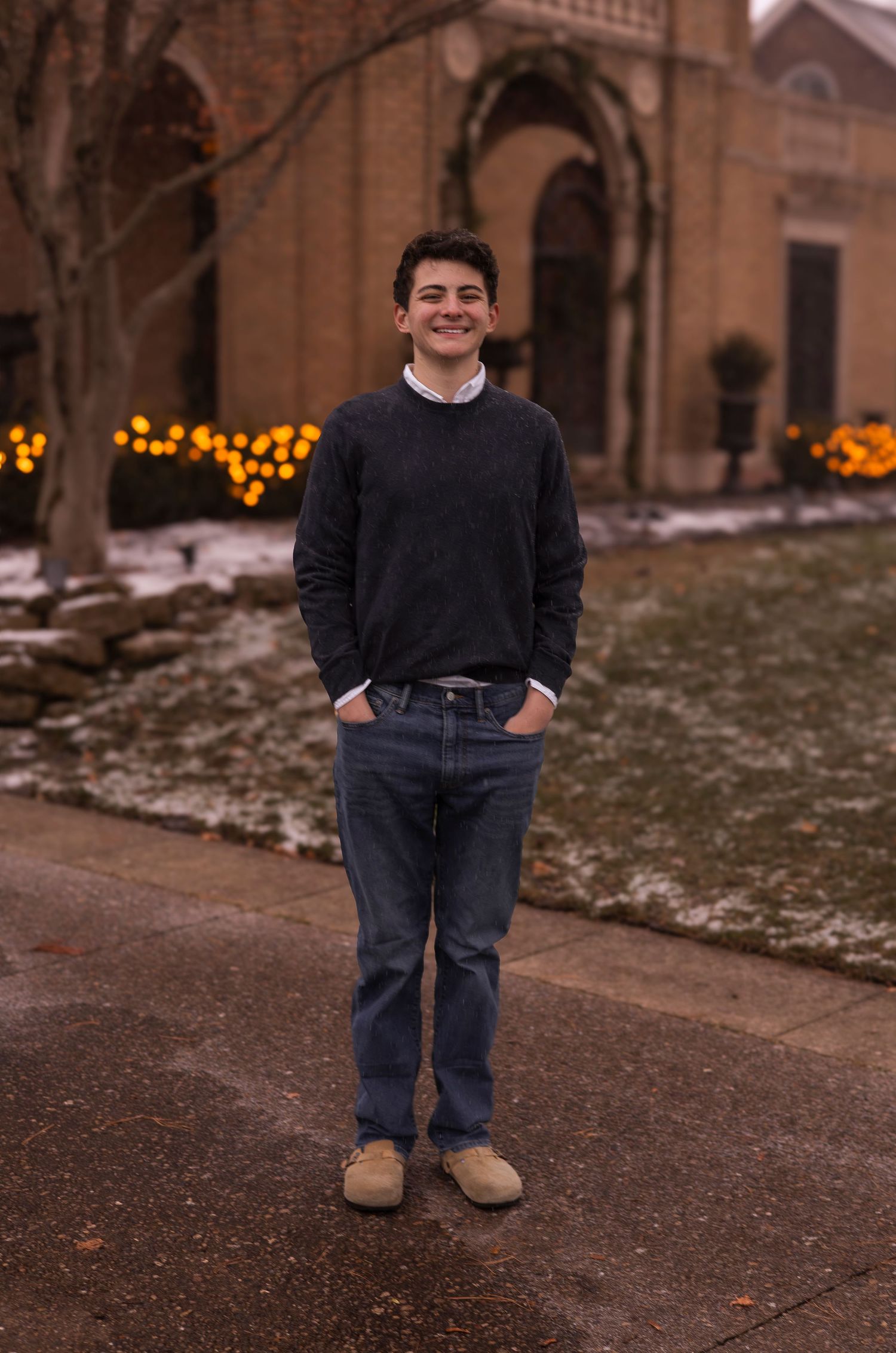 Teenage boy in navy sweater and jeans stands on a snowy sidewalk outside a brick building with lights.