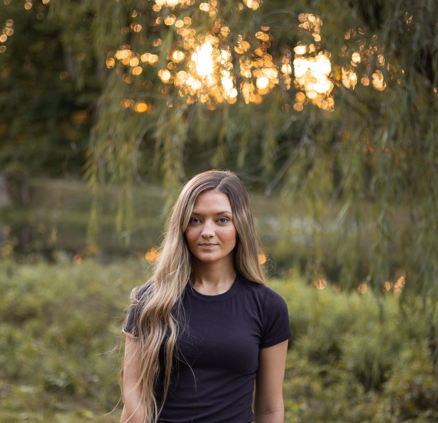 Young blonde woman standing outdoors at golden hour under weeping willow tree.