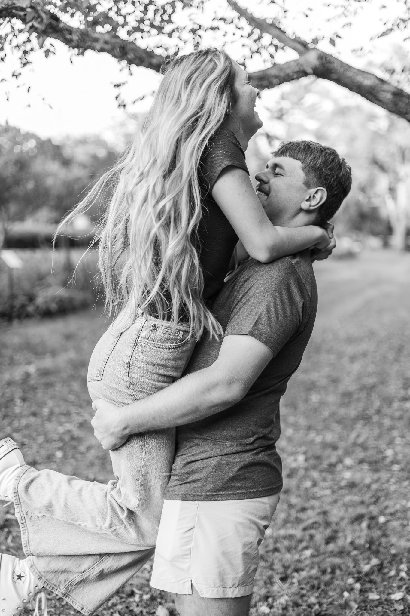 A romantic black and white photo of a young couple embracing and kissing outdoors under a tree.