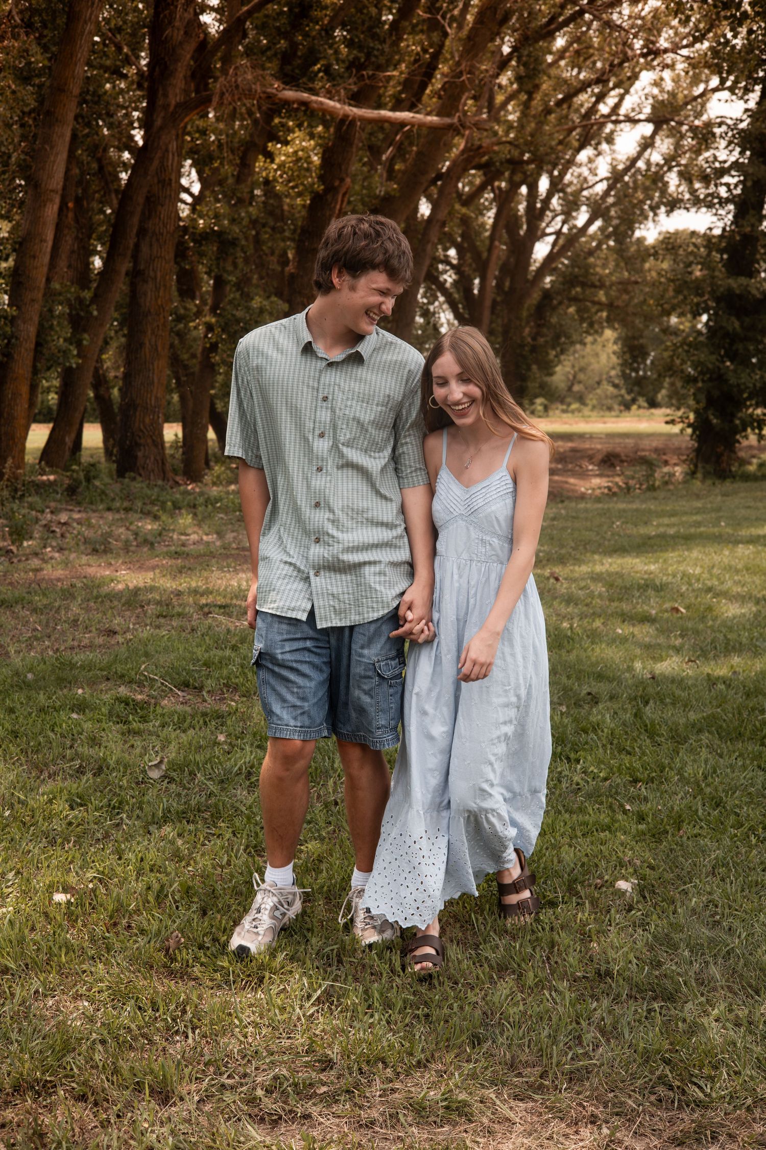 Smiling young couple walking hand in hand through a lush green park with tall trees.