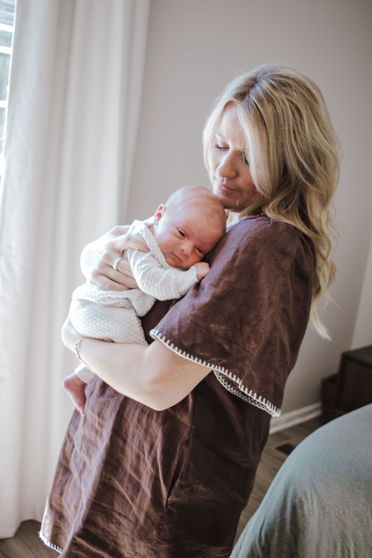 Blonde mother tenderly holding newborn baby in white outfit while wearing a brown linen dress.