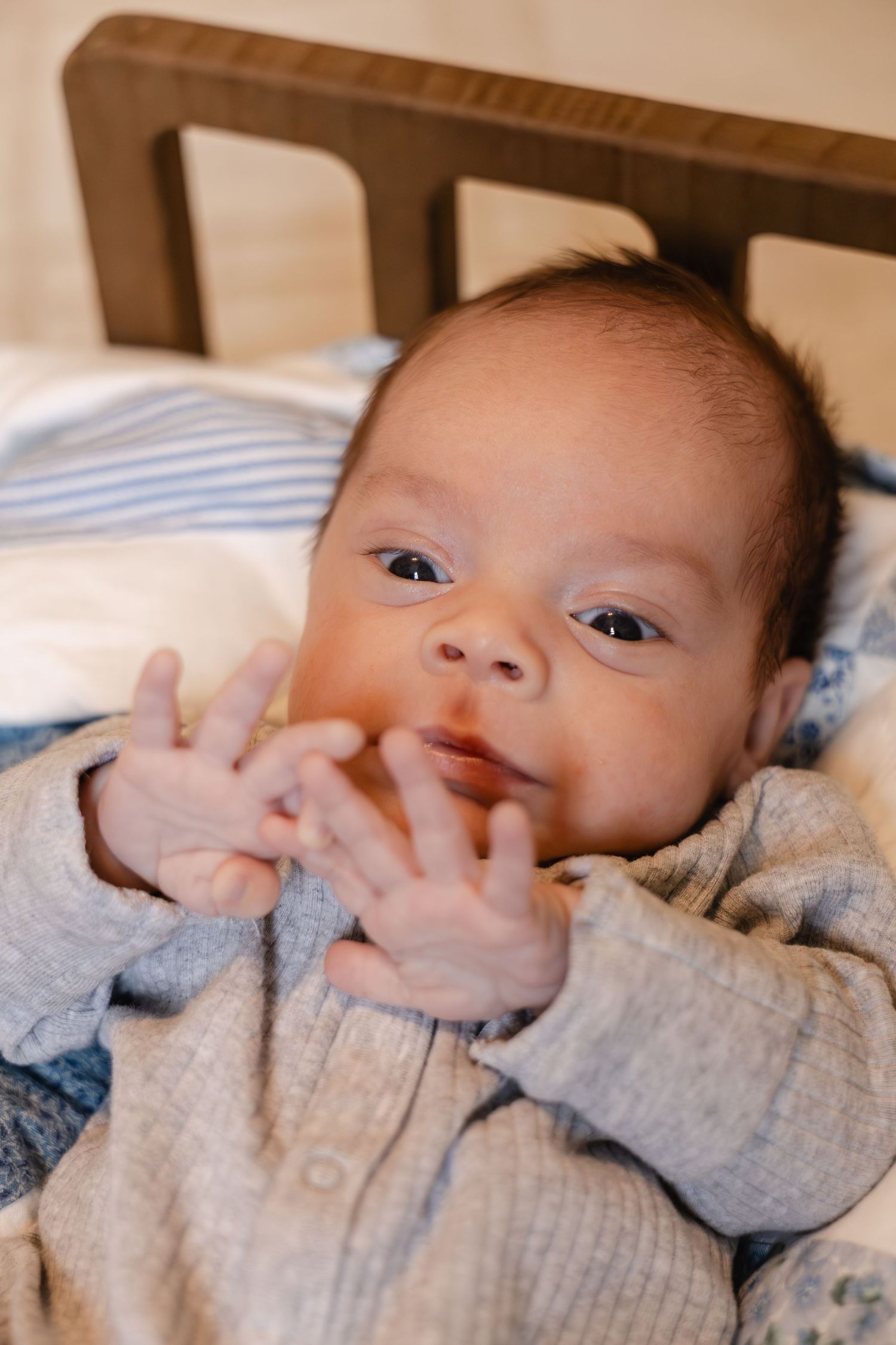Adorable newborn baby in gray onesie lying in wooden crib with hands near face.