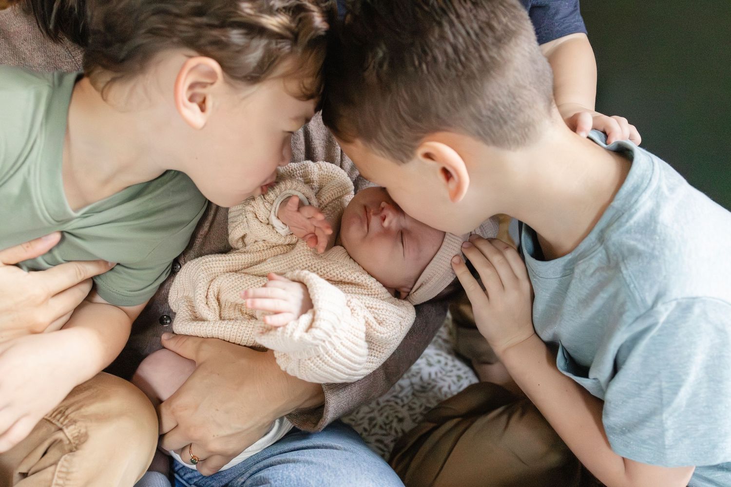 Two siblings lean in to kiss their newborn baby sister held in mothers arms.