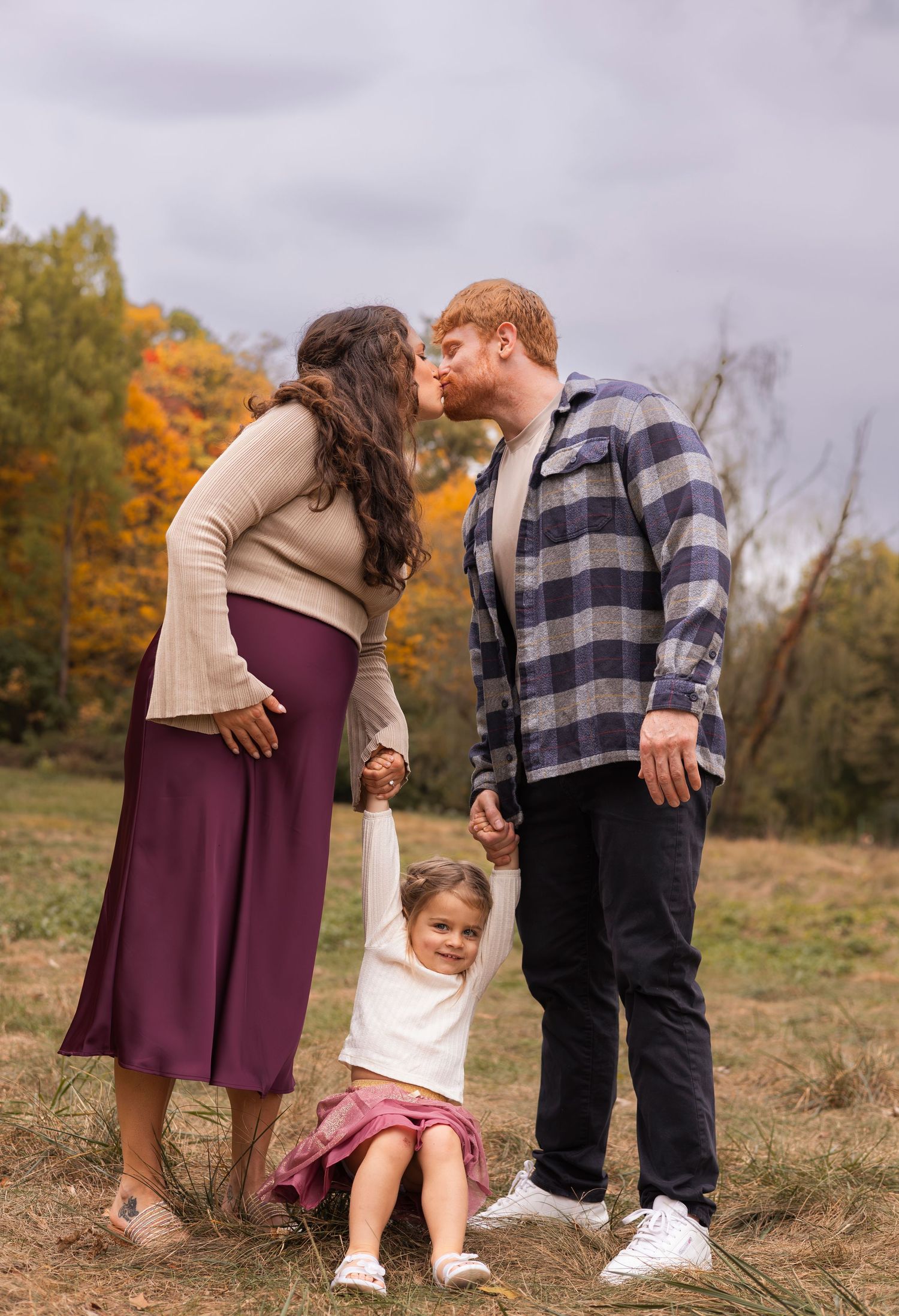 Expecting couple kissing while their young daughter swings between them in autumn field.